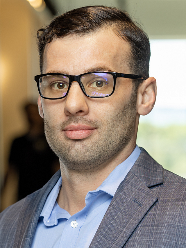 Man wearing glasses, a blue shirt, and gray plaid blazer against a softly blurred background.
