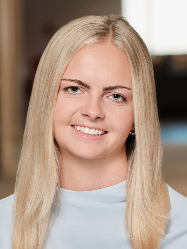 Professional headshot of a smiling woman with long blonde hair wearing a light blue sleeveless top.