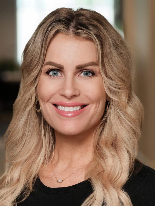 Professional headshot of a smiling woman with long wavy blonde hair wearing a black top.