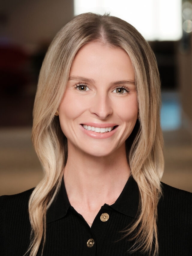 Professional headshot of a smiling woman with straight blonde hair wearing a black collared top.