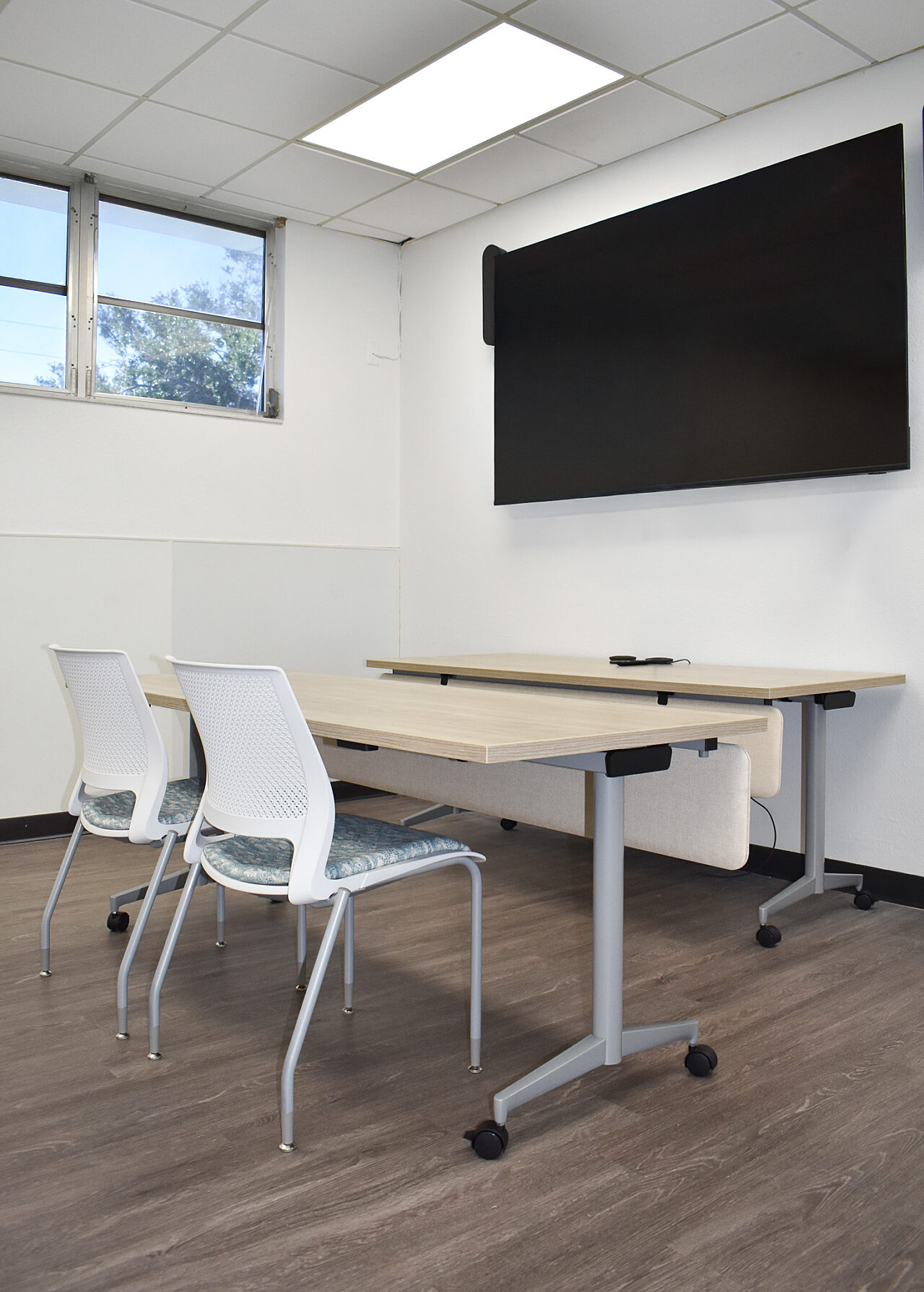 Small meeting room with two white chairs, rolling tables, and a large wall-mounted screen.