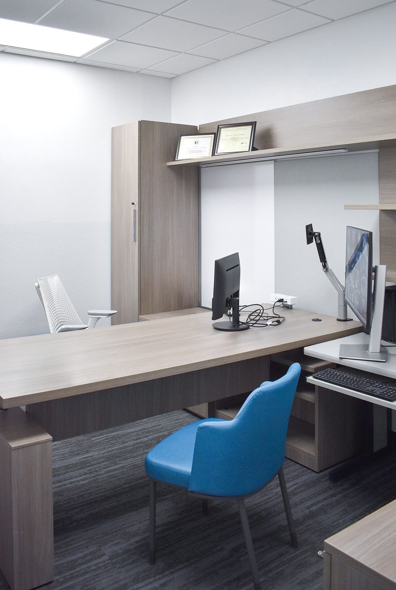 Office with wood desk, dual monitors, storage cabinets, and a blue guest chair.
