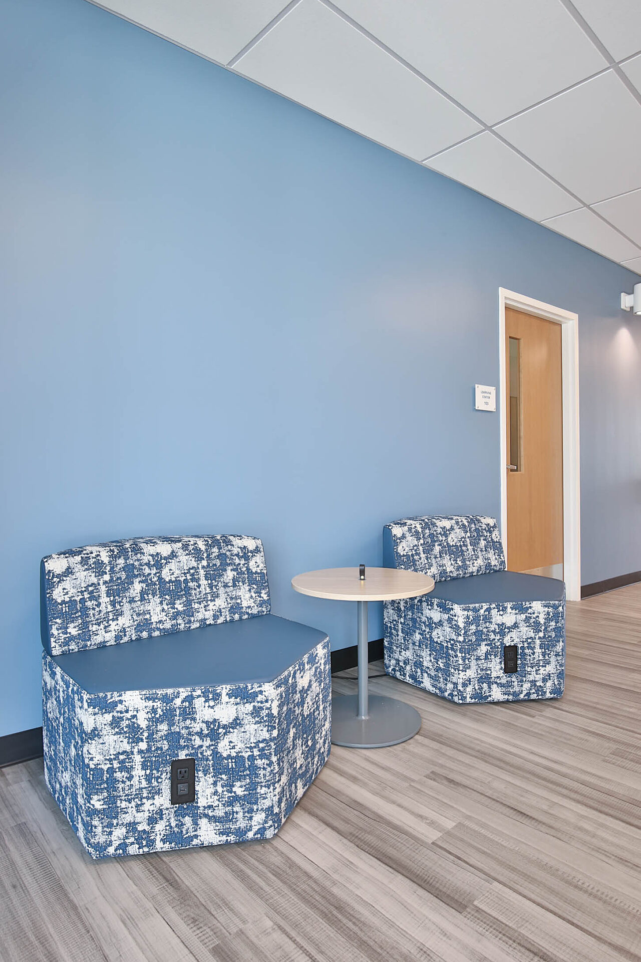 Lounge area with two blue-and-white patterned chairs, a round table, blue accent wall, and wood-look flooring.