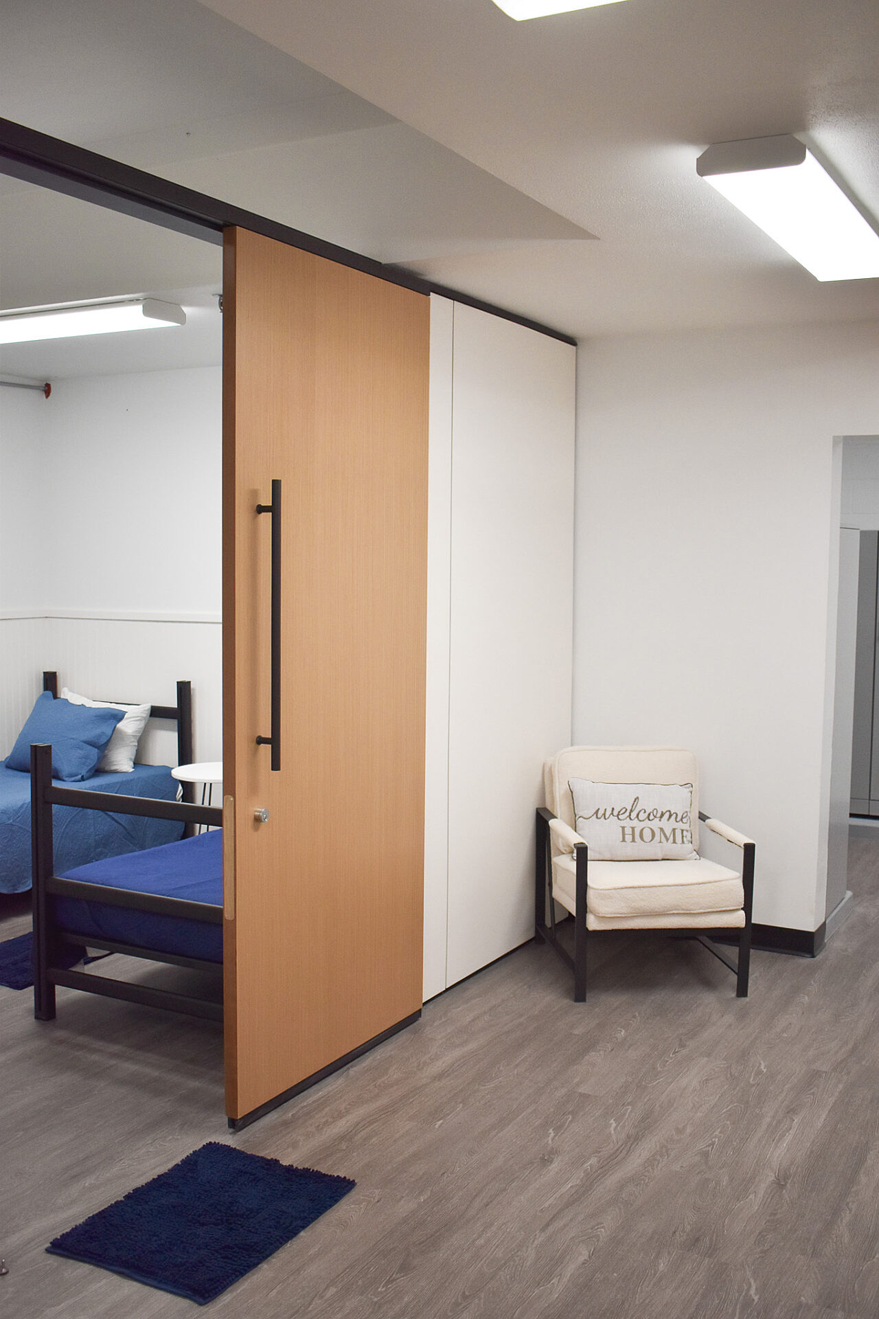 Room with bunk beds, a sliding wooden door, and a white cushioned chair on a gray floor.