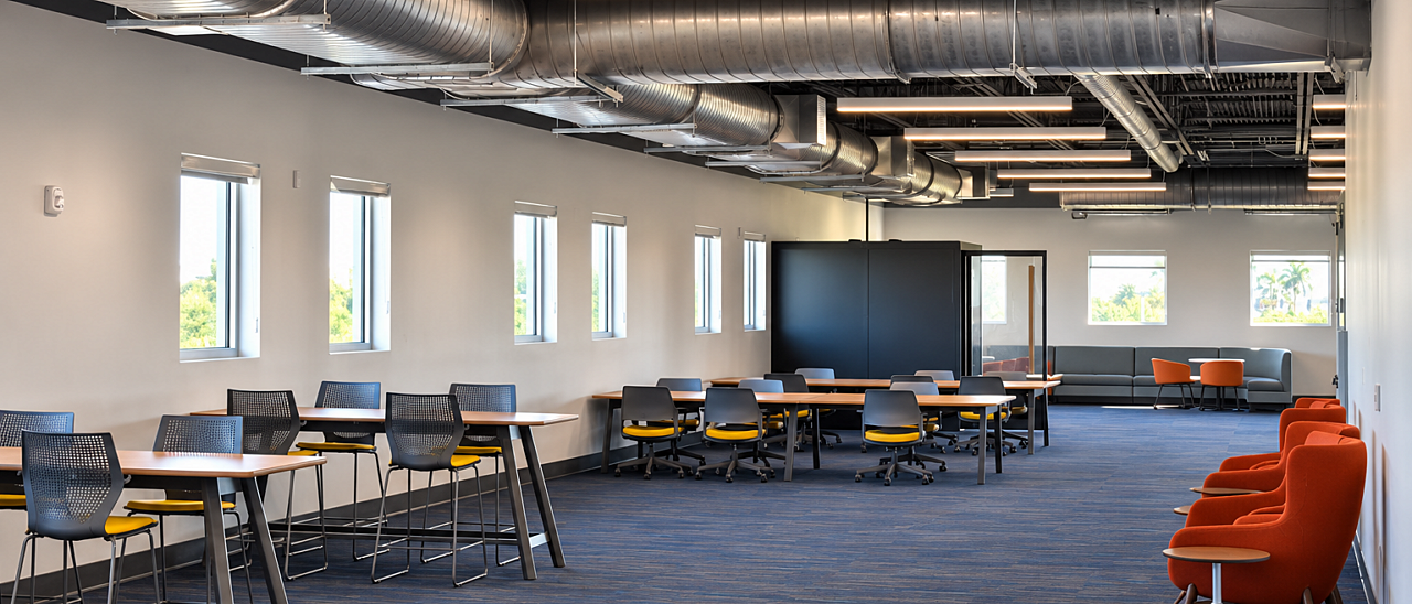 Library study area with communal tables, black and yellow chairs, orange lounge seating, and exposed ductwork.