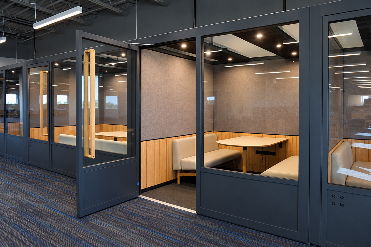 Glass-enclosed library study pods with cream bench seating, wood tables, and acoustic wall panels.