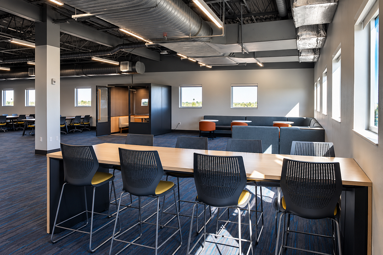 Library study area with a high-top communal table, black chairs, lounge seating, and a private study pod.