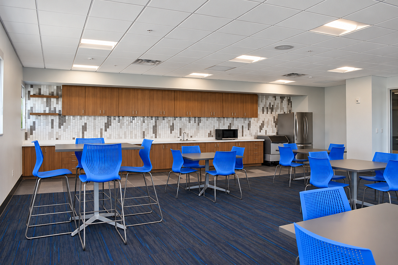 Modern library breakroom with blue chairs, wood cabinetry, café tables, and patterned tile backsplash.