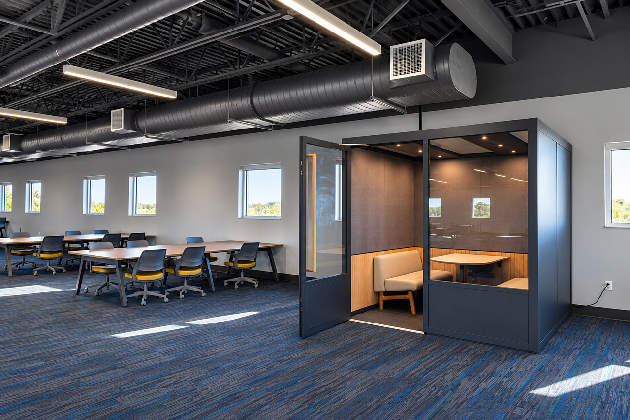 Library study area with communal tables, black and yellow chairs, and an enclosed glass study pod.