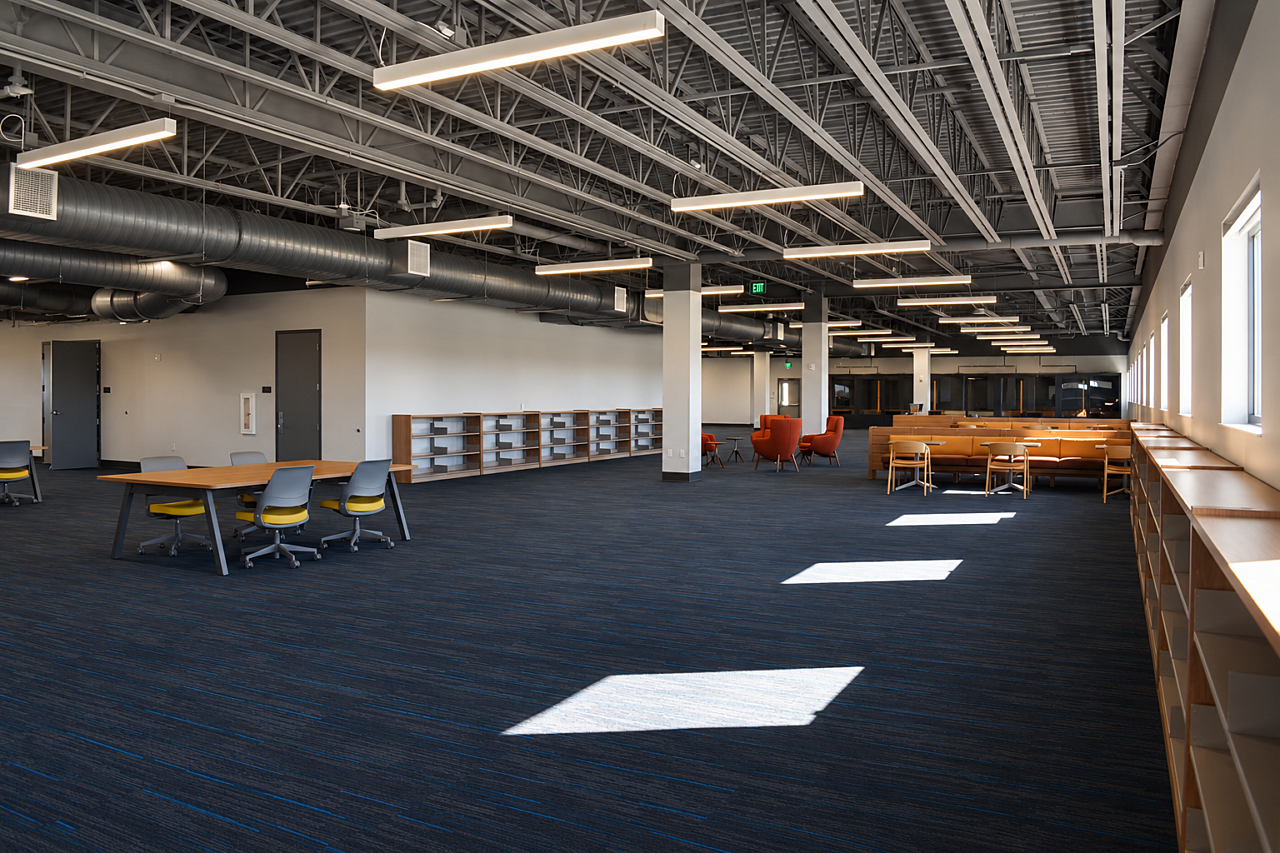 Spacious library with exposed ceilings, blue carpeting, bookshelves, and collaborative seating areas.
