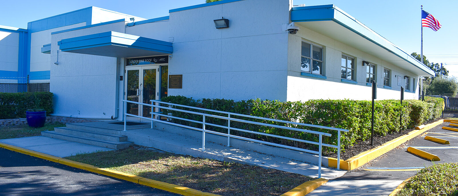 Single-story white building with a ramp entrance, blue trim, hedges, and a U.S. flag outside.
