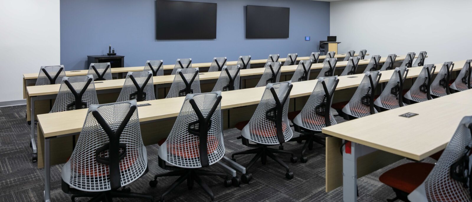 Training room with rows of tables, ergonomic seating, dual display screens and presentation area.