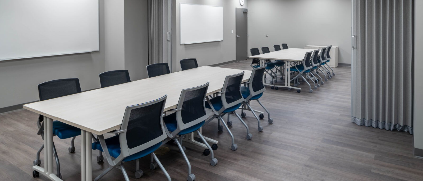 Two connected meeting rooms with long tables, rolling chairs, whiteboards, and gray partition curtains.