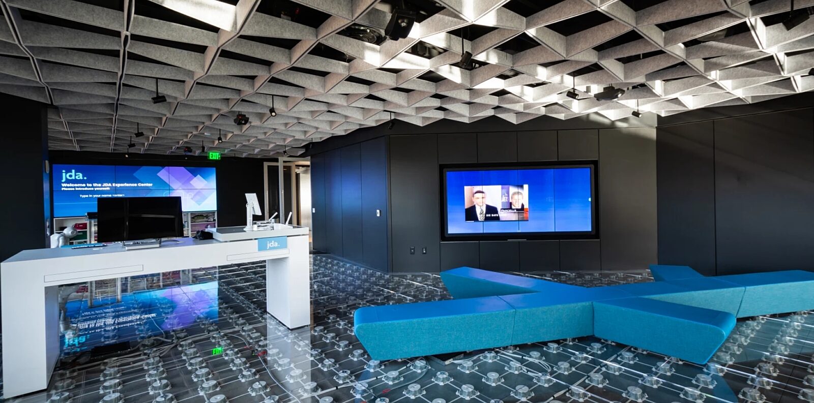 Modern experience center with geometric acoustic ceiling, digital displays, and blue modular seating on a glass-raised floor system.