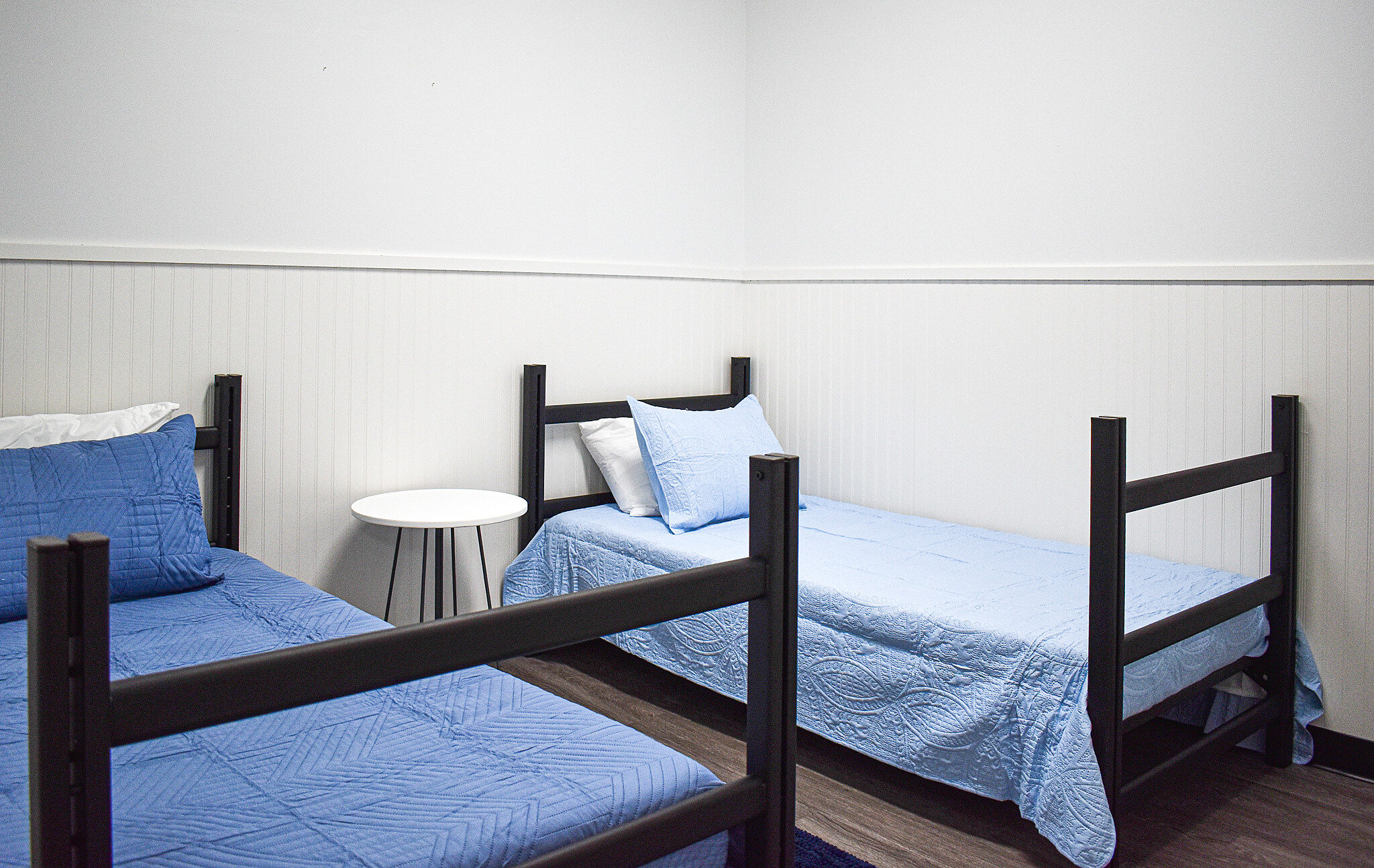 Two twin beds with blue bedding and a small white side table in a simple, light-colored room.