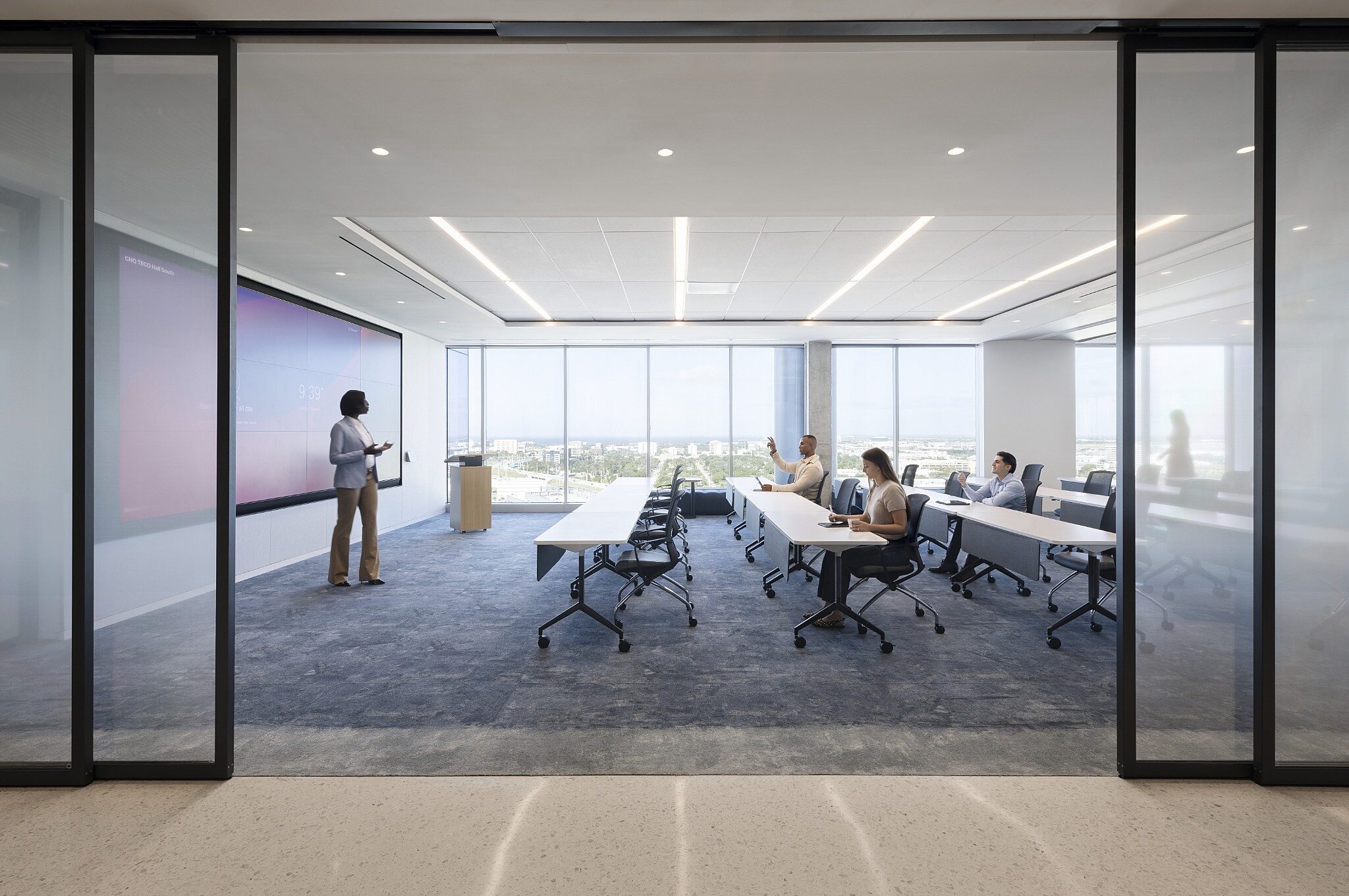 Modern training room with tables, rolling chairs, and large display, framed by glass walls and floor-to-ceiling windows.