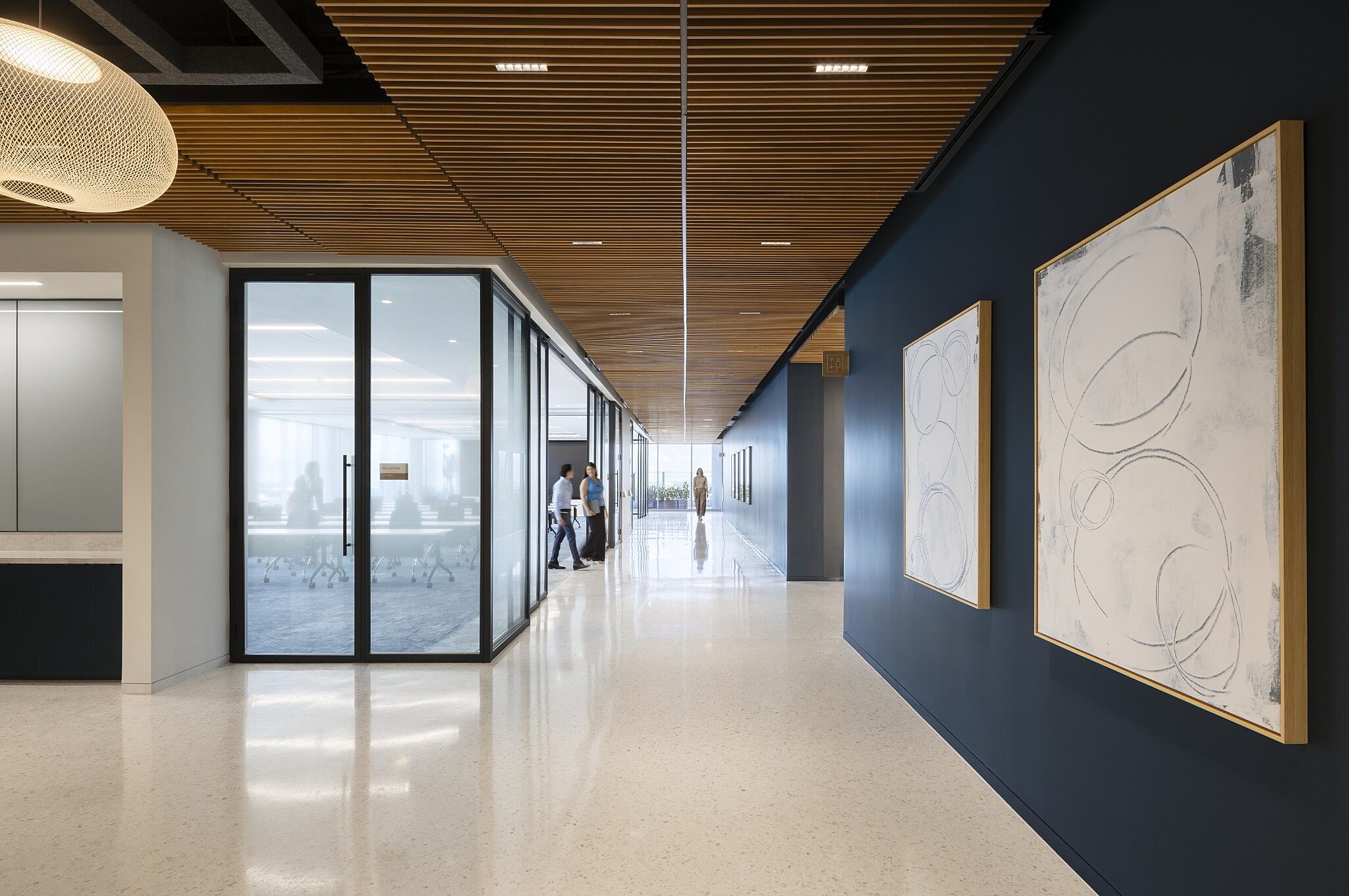 Modern office corridor with wood slat ceiling, glass-walled meeting rooms, and large abstract artwork along a dark accent wall.