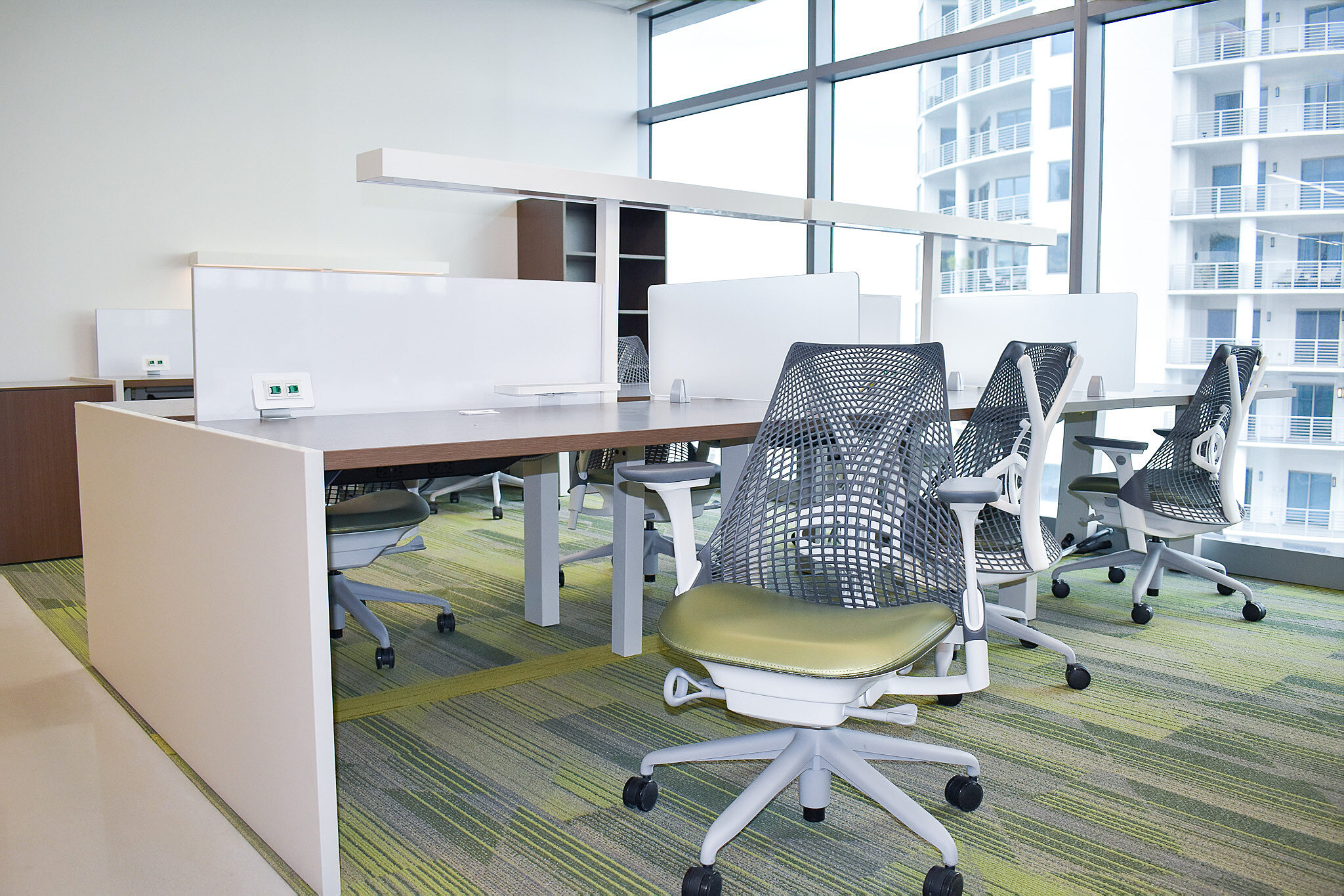 Benching workstation with ergonomic mesh chairs and privacy screens near floor-to-ceiling windows.