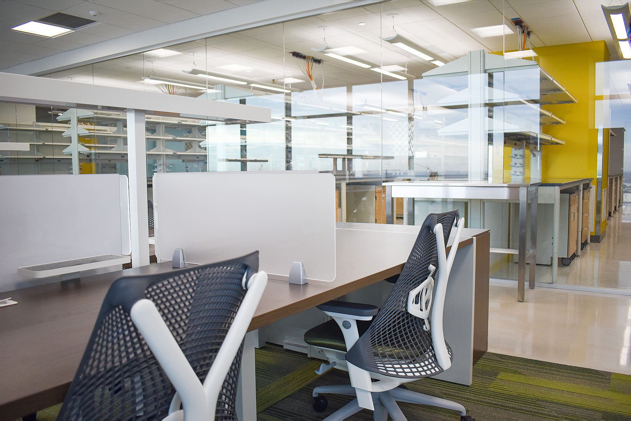 Modern workstation with privacy panels and ergonomic chairs beside glass-enclosed lab space.