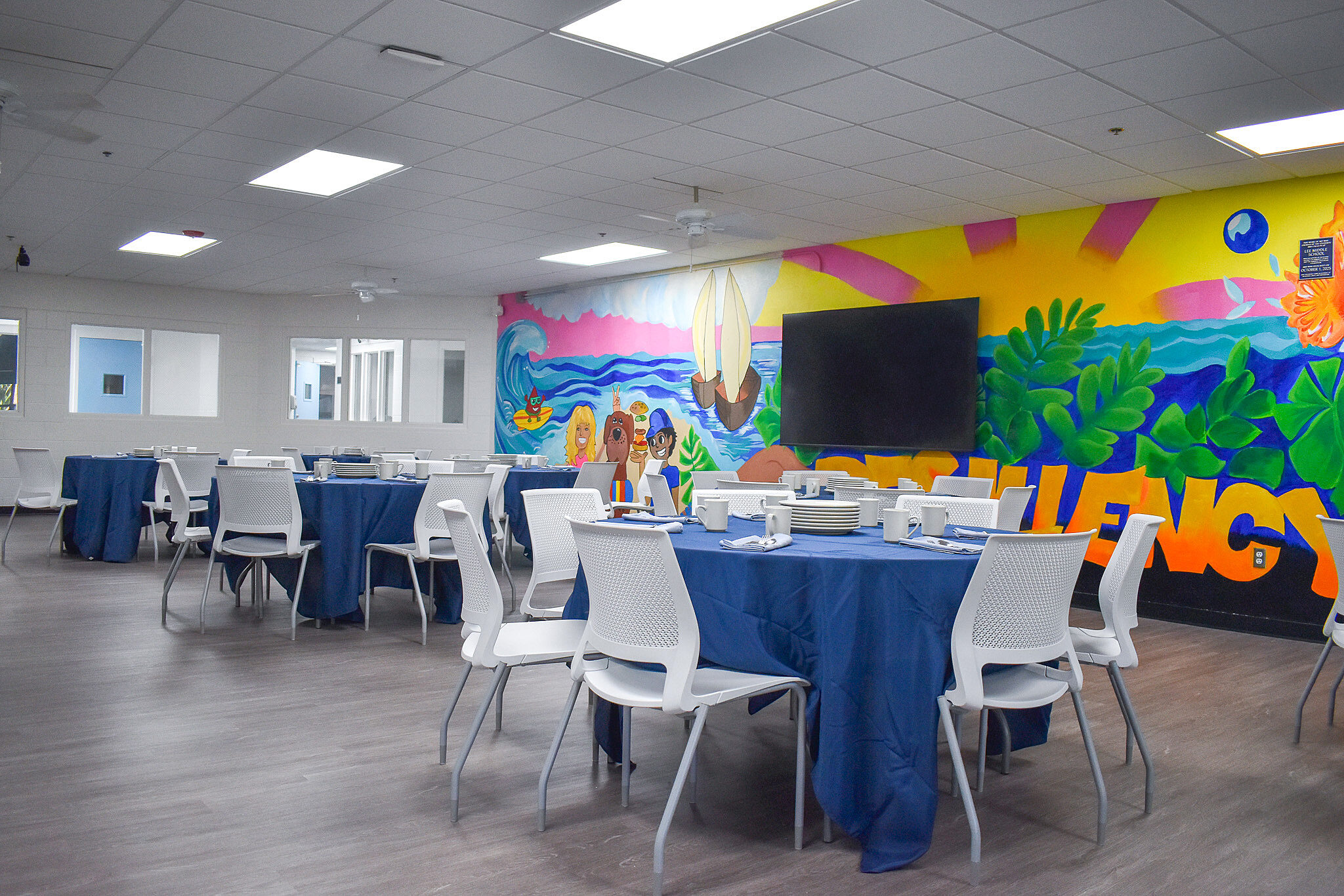 Bright community room with round tables, white chairs, and a colorful mural reading “Resiliency.”
