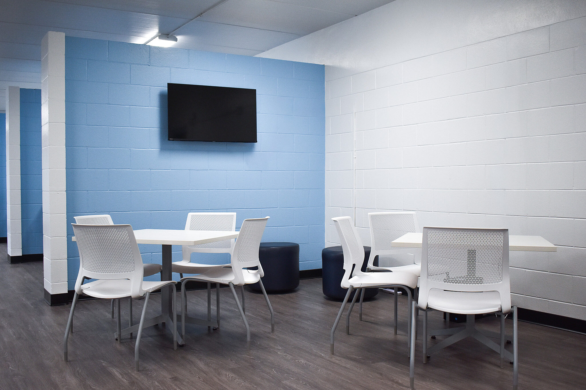 Break area with white tables and chairs, blue accent wall, TV, and dark wood-style flooring.