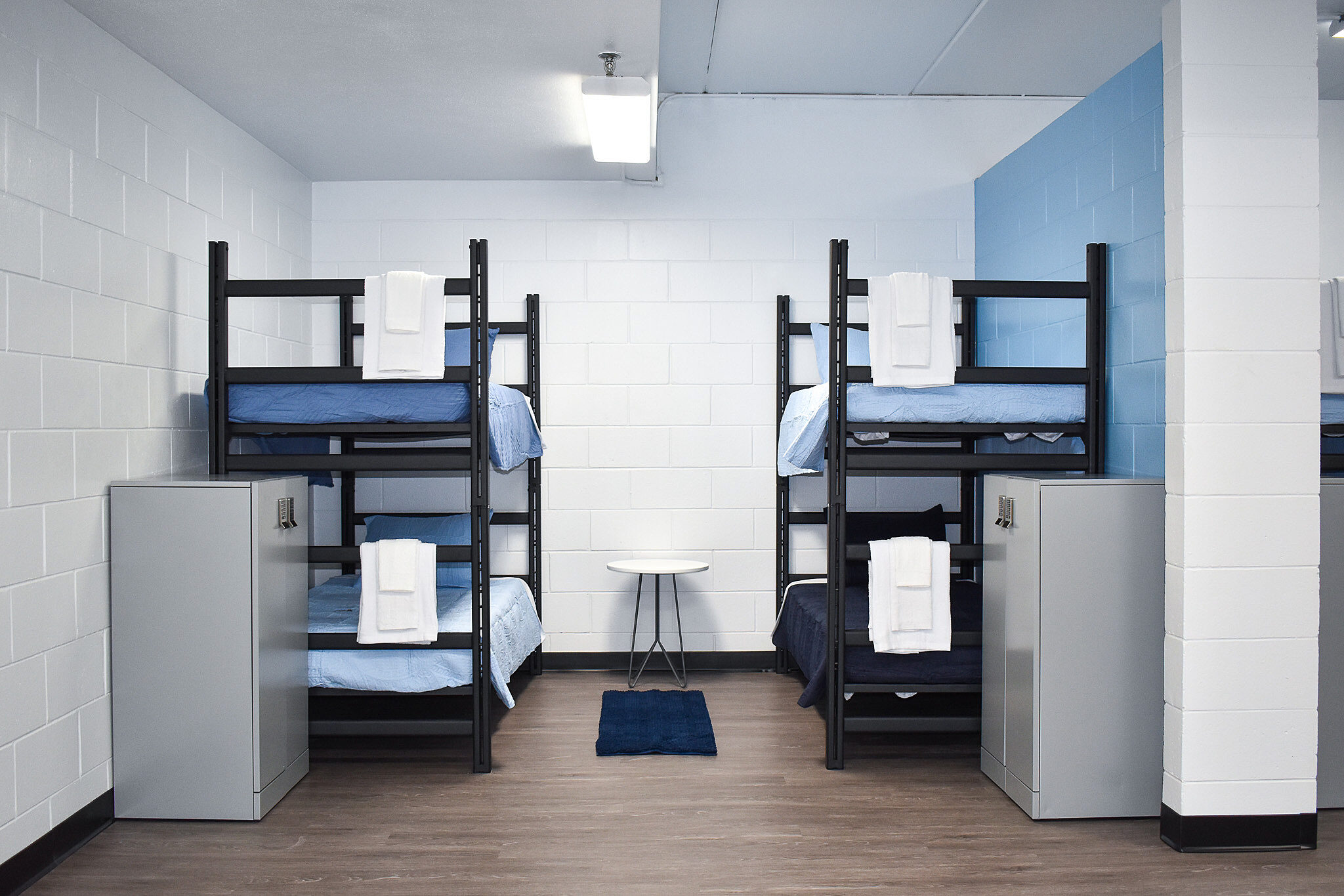 Two sets of bunk beds with blue bedding, metal lockers, and a small table in a clean dorm room.