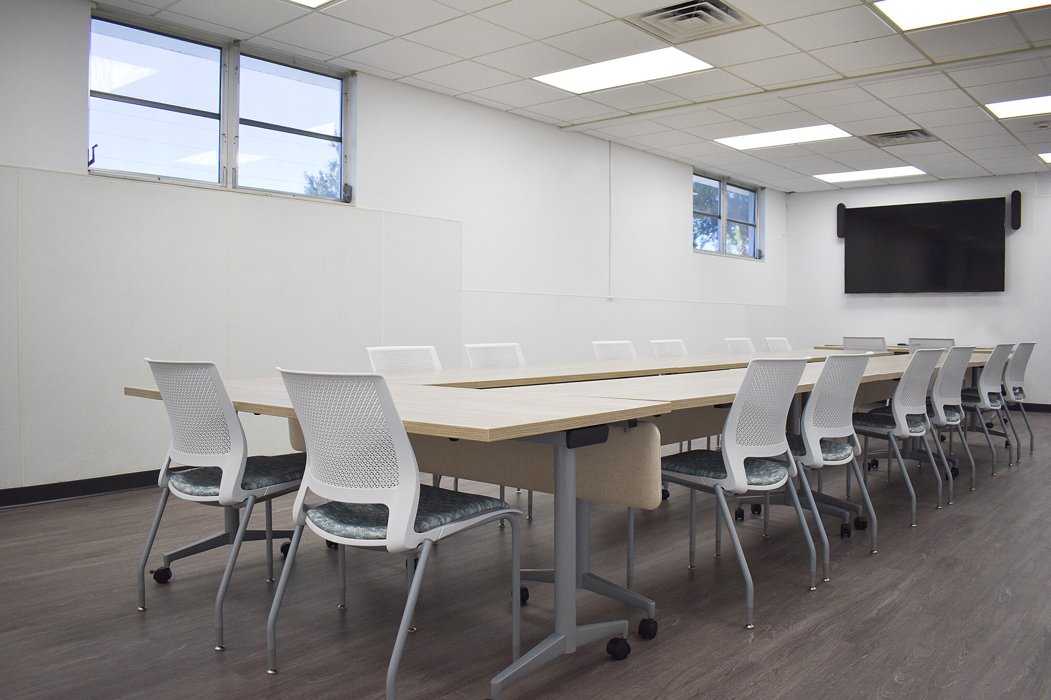 Large meeting room with U-shaped tables, white chairs, wood floors, and a wall-mounted TV.