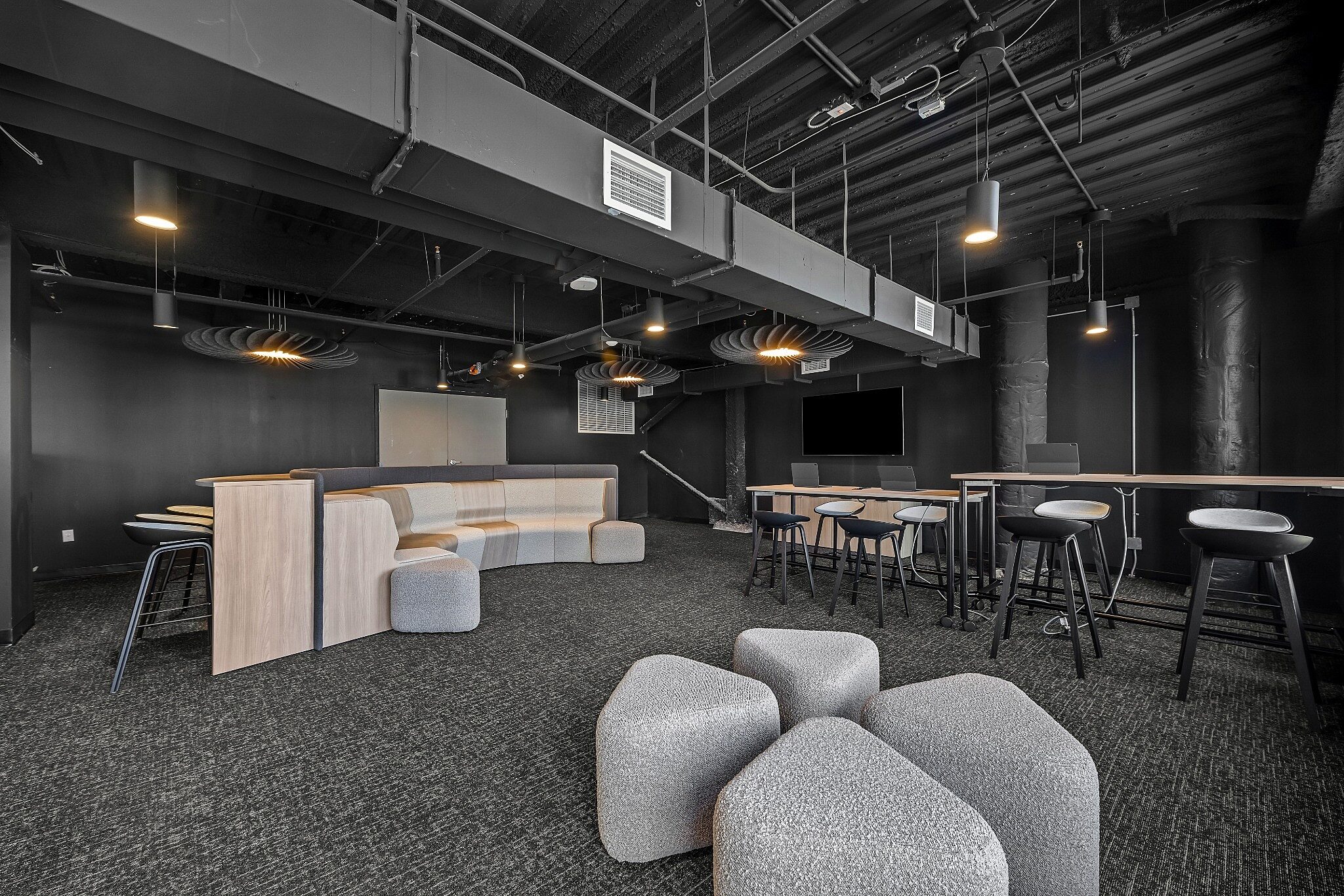 Modern lounge workspace with modular seating, high-top tables, and exposed ceiling elements in a dark, contemporary interior.