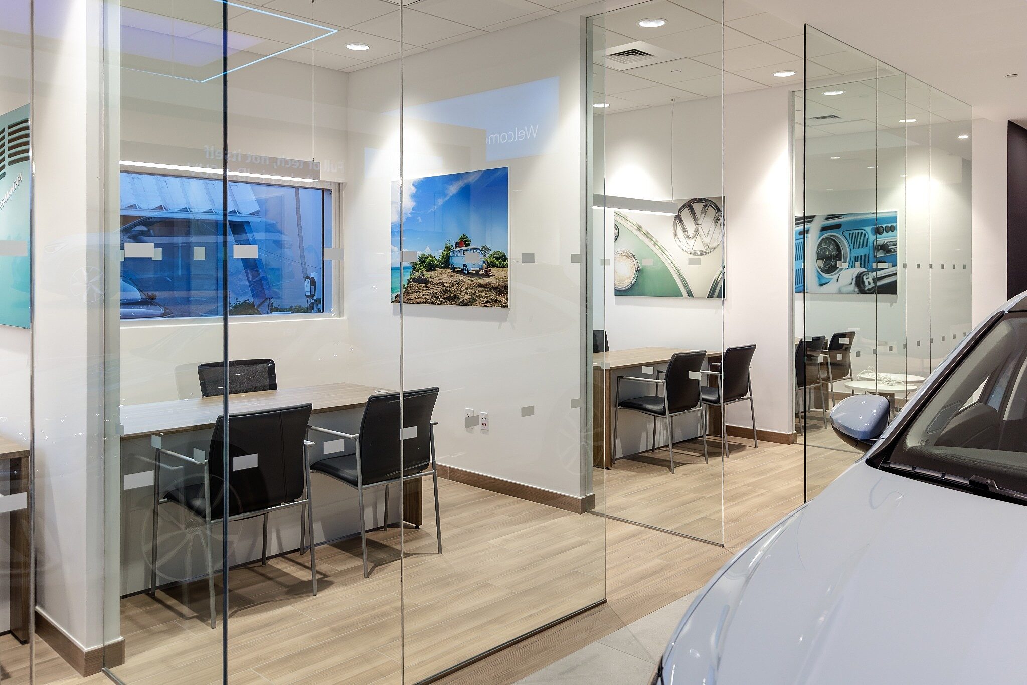 Glass-enclosed meeting rooms with desks and chairs, featuring automotive-themed artwork and a vehicle partially visible in a modern showroom setting.