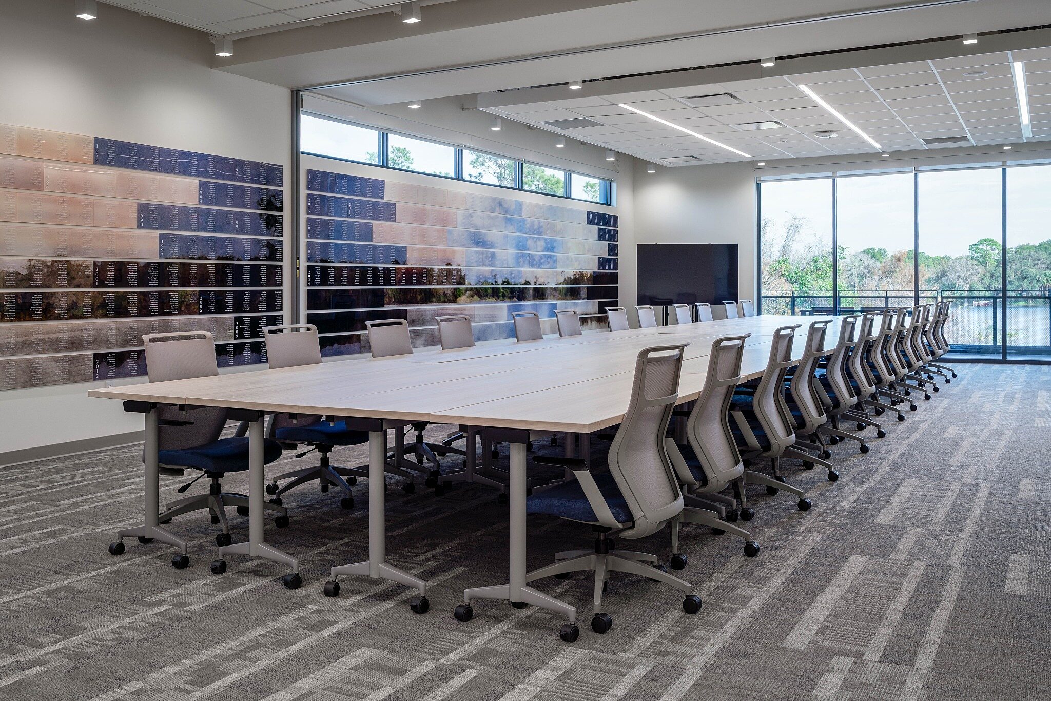 Modern conference room with long table, rolling chairs, large windows, and feature wall of material panels.
