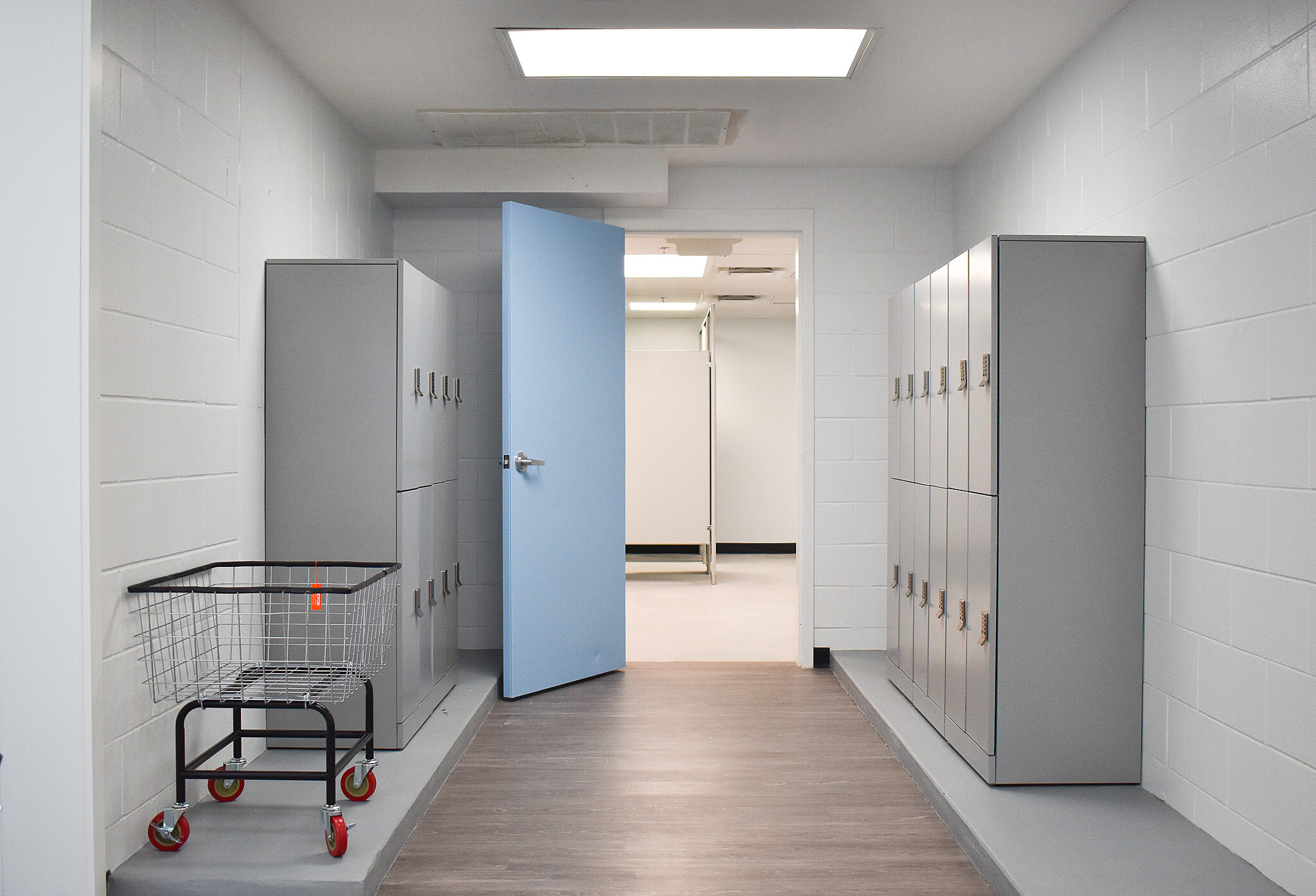 Locker room with gray lockers, rolling laundry cart, blue door, and bright overhead lighting.