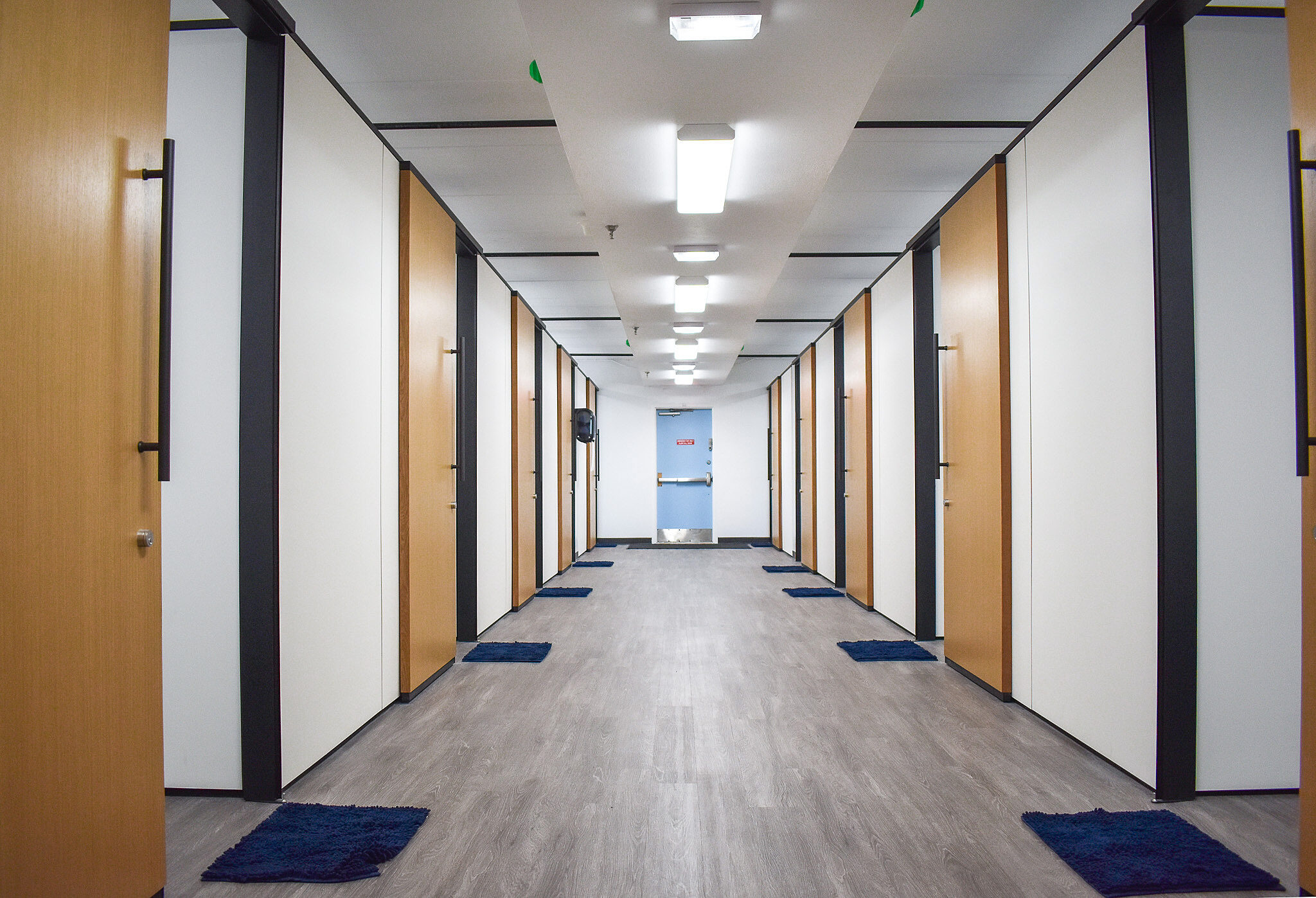 Straight hallway lined with wood doors, white walls, blue floor mats, and overhead lighting.