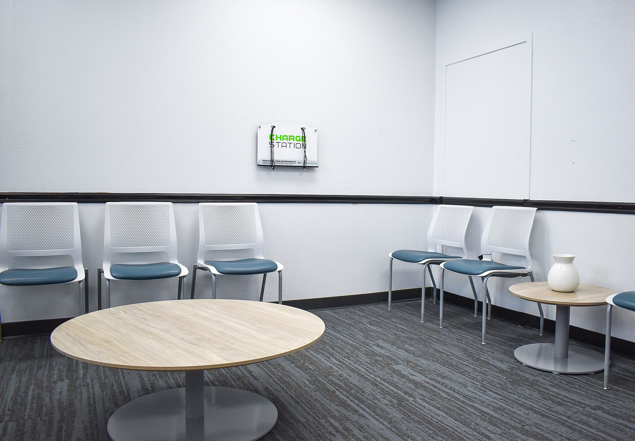 Simple waiting room with white chairs, round tables, gray carpet, and a wall-mounted brochure rack.
