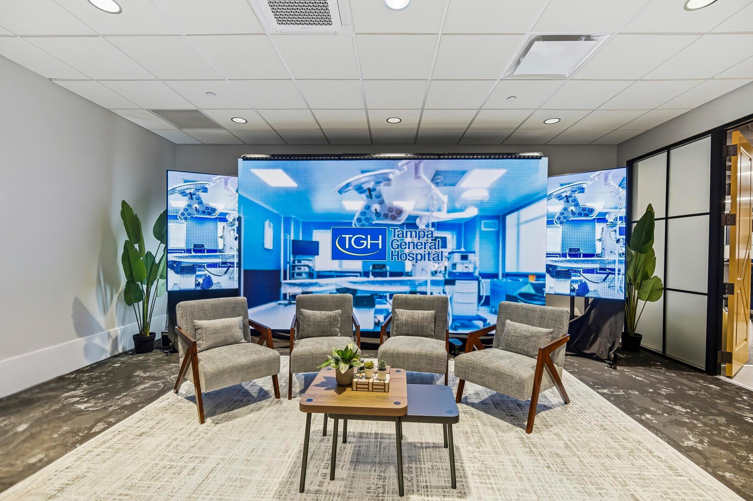 Lounge seating area with four chairs and coffee tables facing a large digital display wall showing Tampa General Hospital branding.