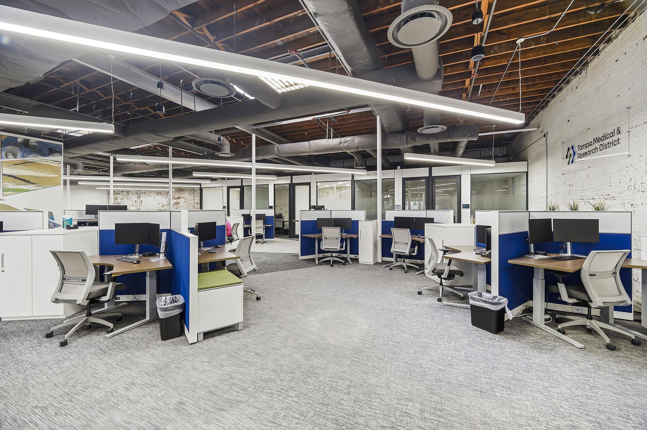 Open office with blue-and-white cubicles, task chairs, and exposed industrial ceiling lighting.