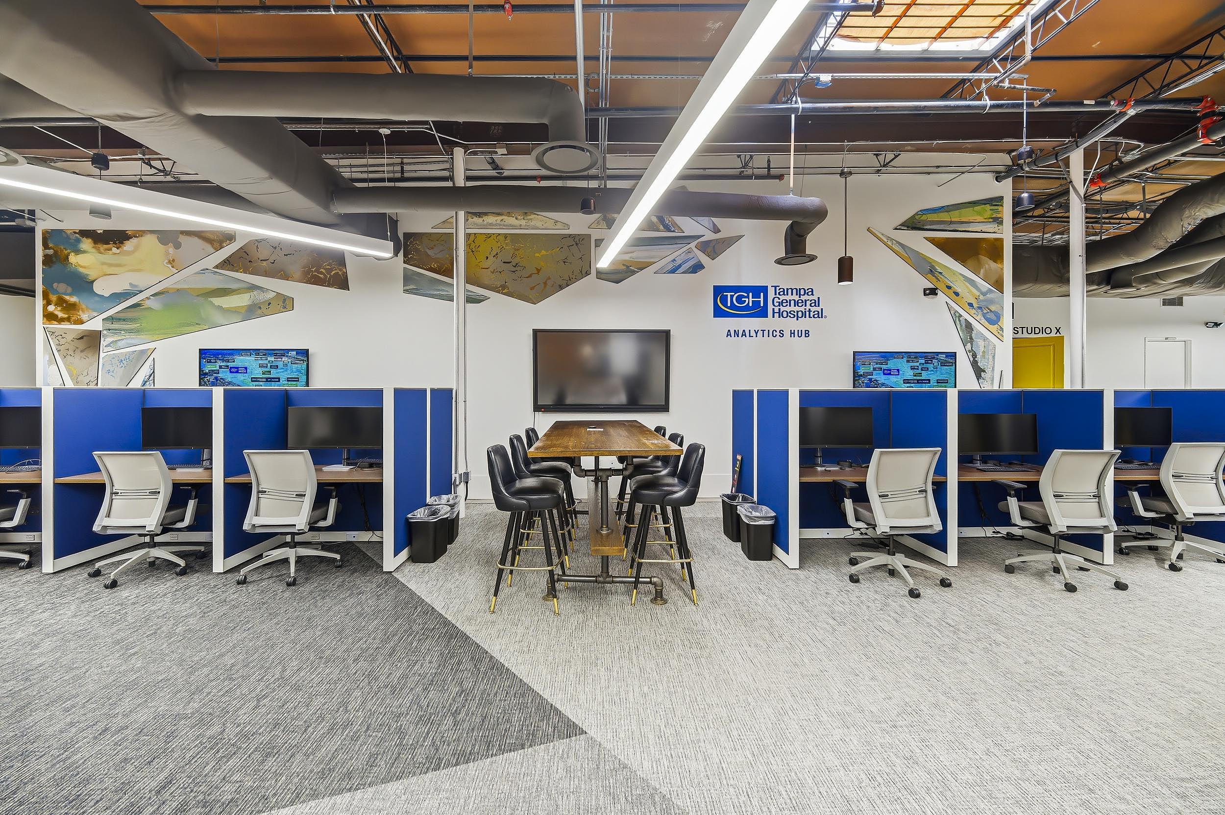 Collaborative workspace with blue cubicles, central meeting table, and Tampa General Hospital Analytics Hub sign.