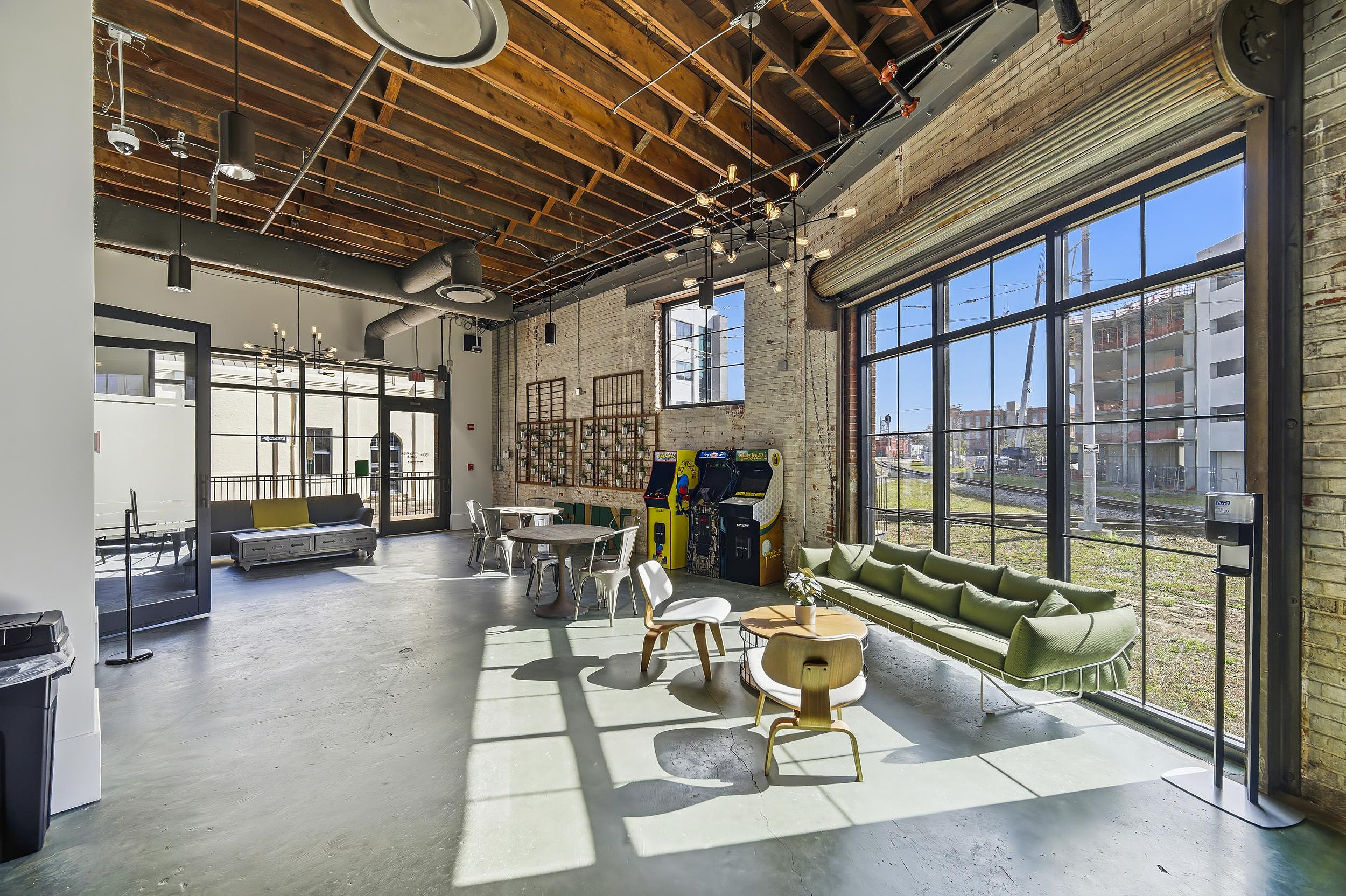 Industrial lounge with large windows, arcade games, green sofa seating, and exposed wood ceiling beams.