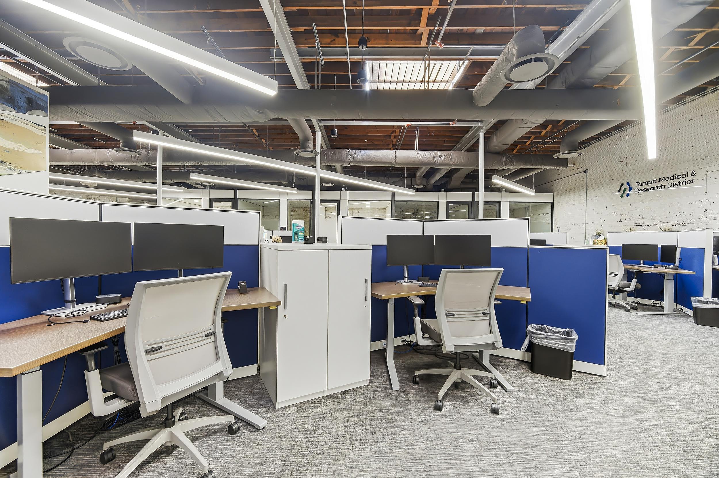 Office cubicles with dual monitors, task chairs, and exposed ceiling with linear lighting.