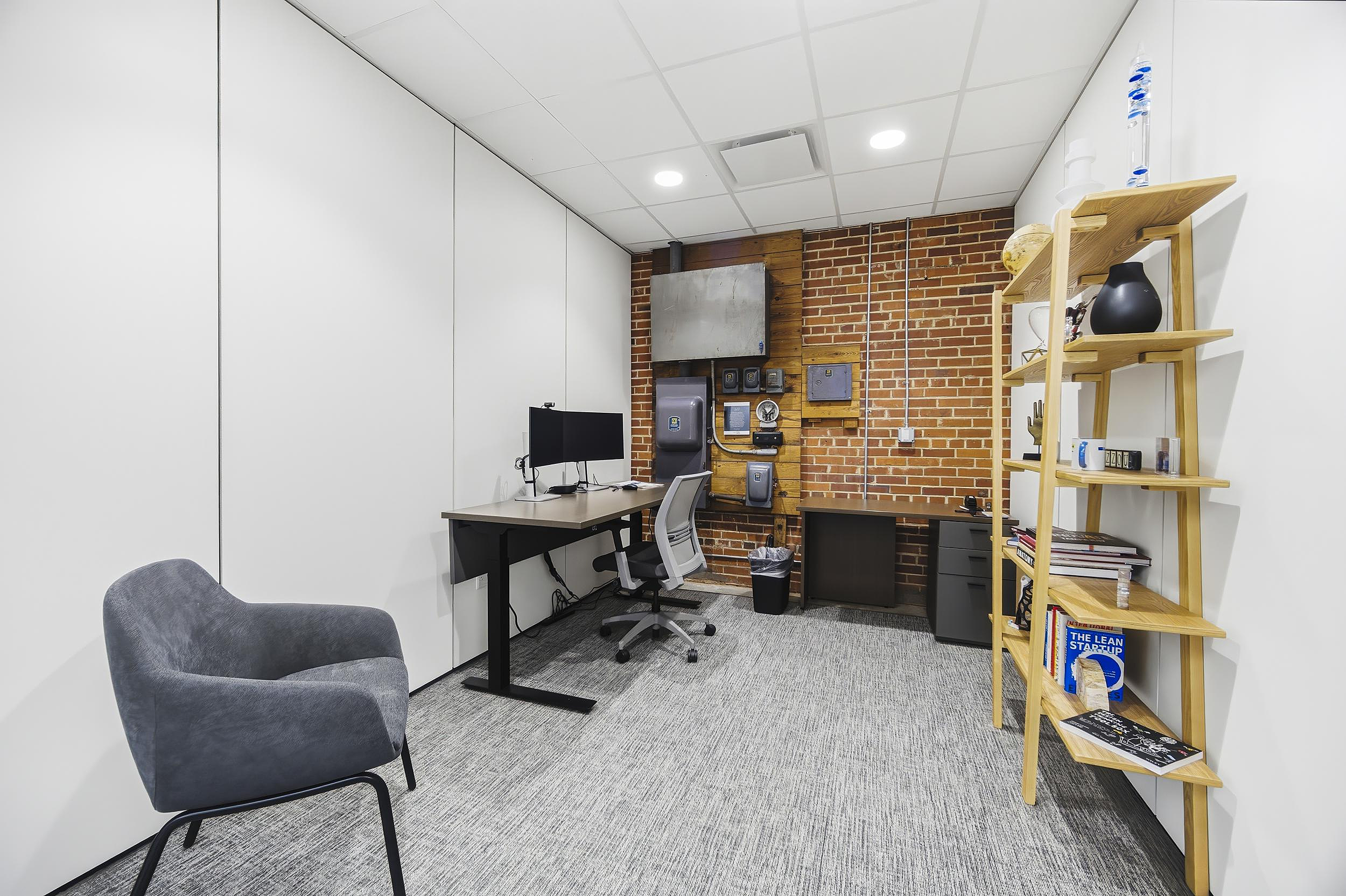 Private office with sit-stand desk, exposed brick wall, shelving, and lounge chair.
