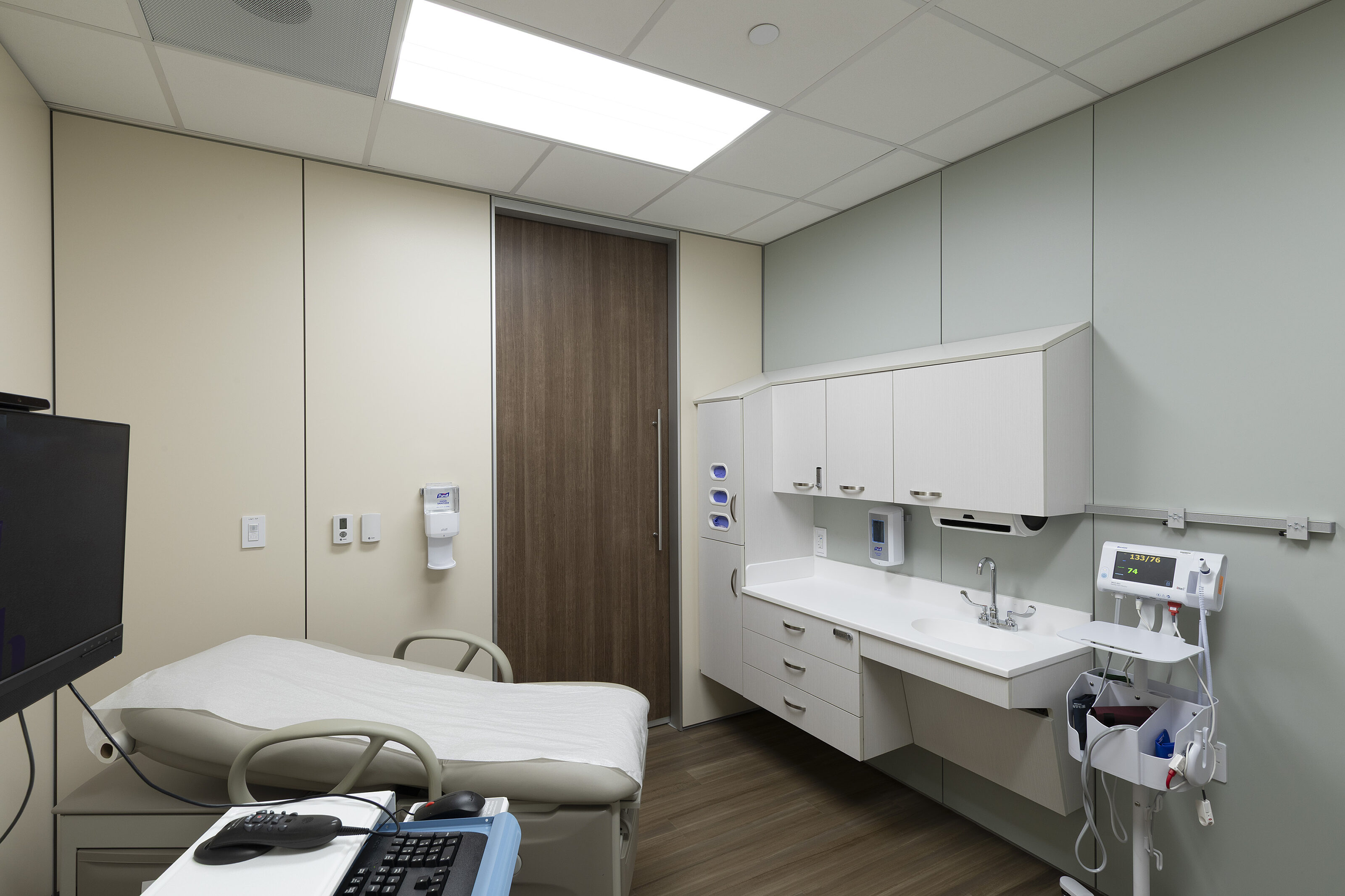 Exam room with patient bed, wall cabinets, sink, medical equipment, and soft neutral finishes.