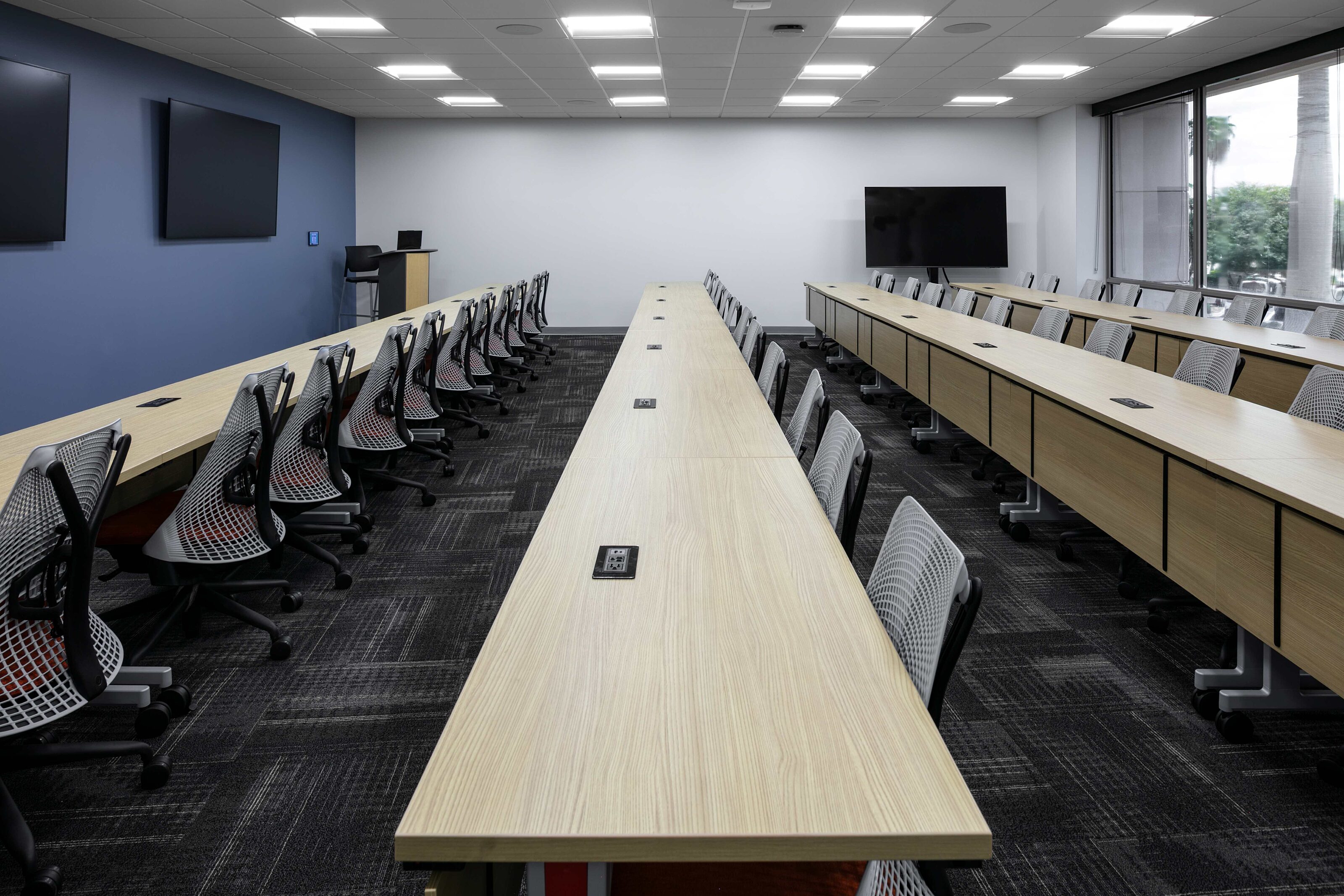 Large training room with rows of light wood tables, mesh chairs and multiple display screens.