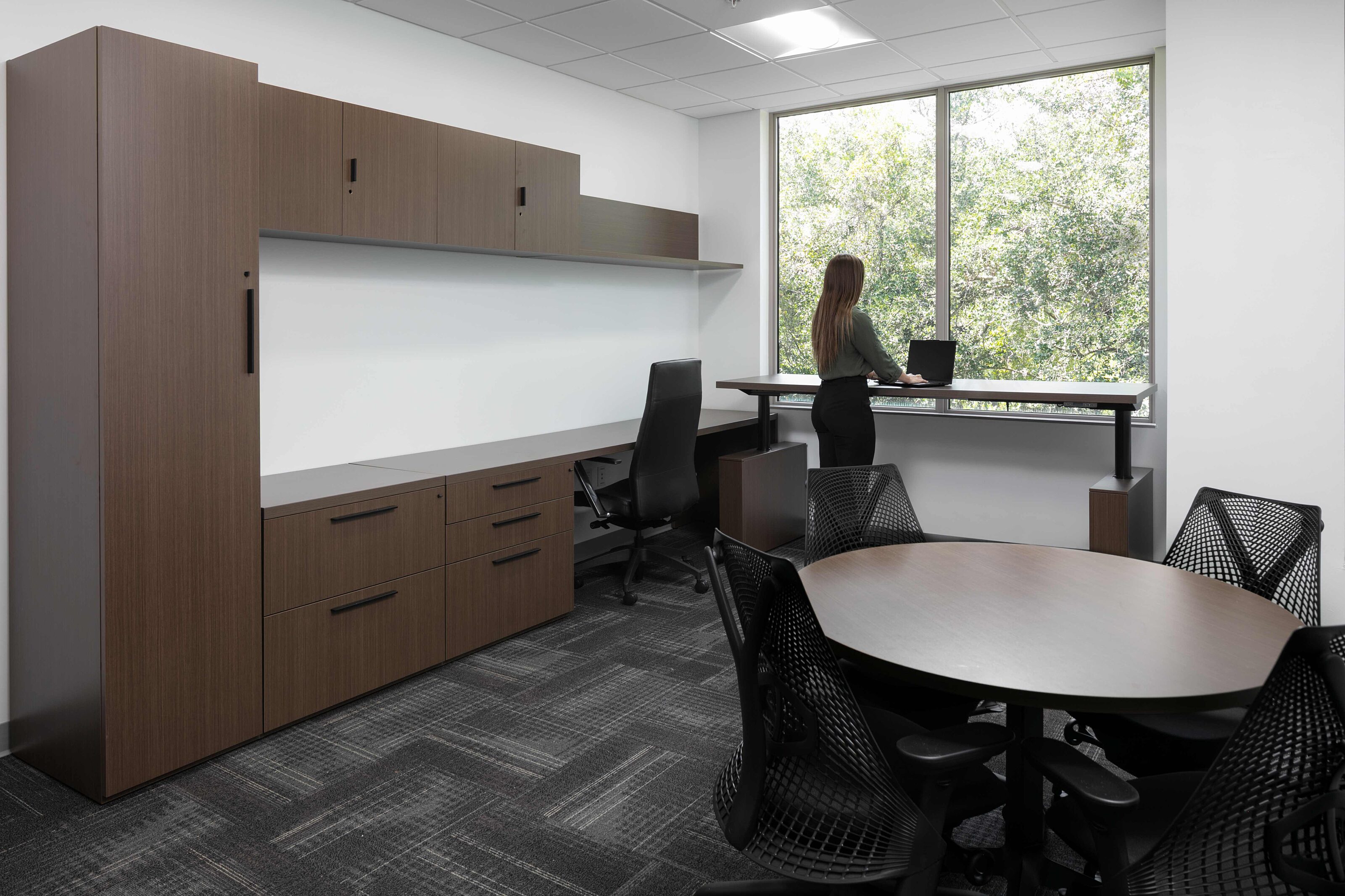Private office with wood storage, meeting table, sit-to-stand desk and large windows.