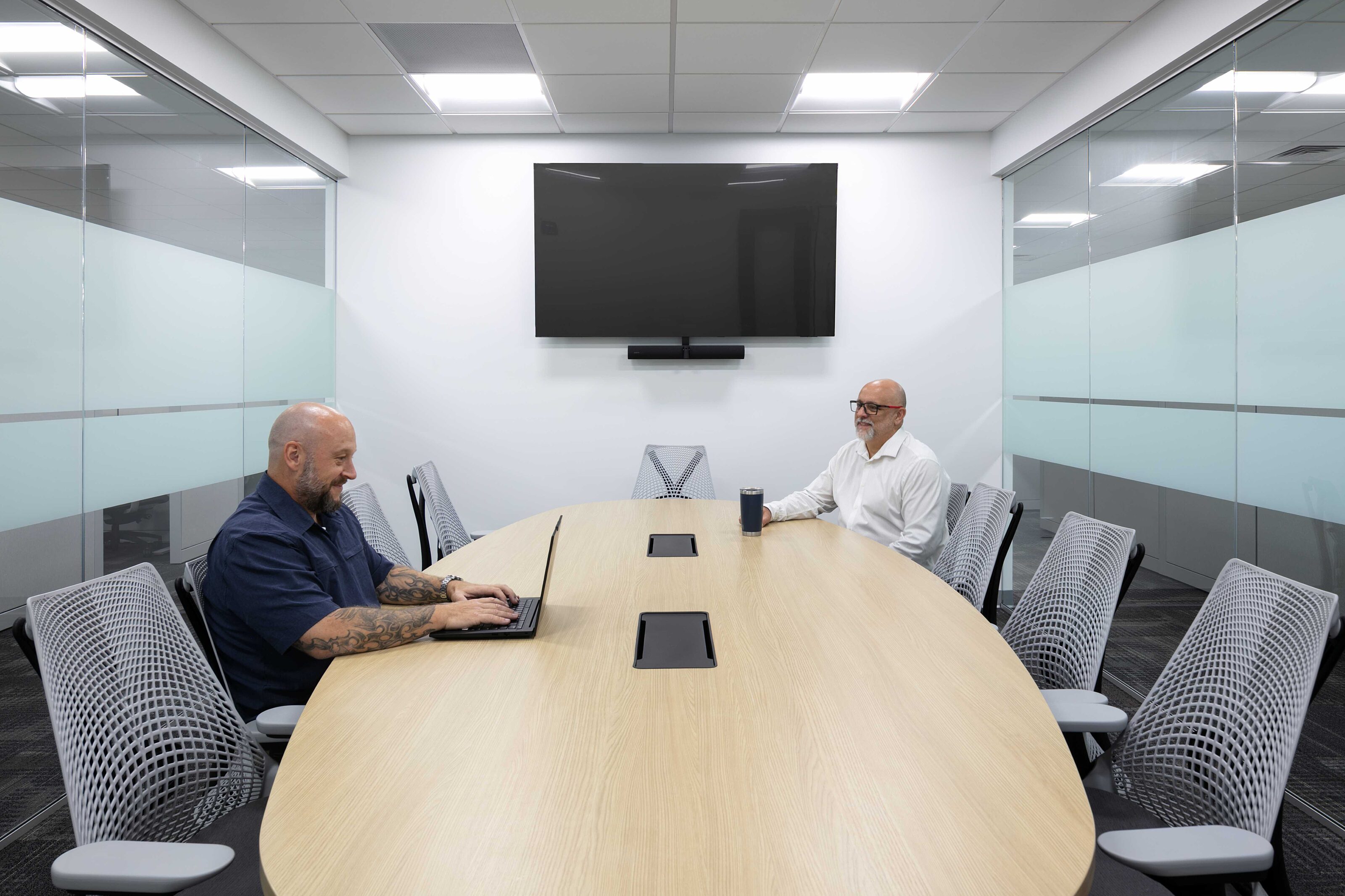 Glass-enclosed conference room with a wood table, mesh chairs and two employees working.