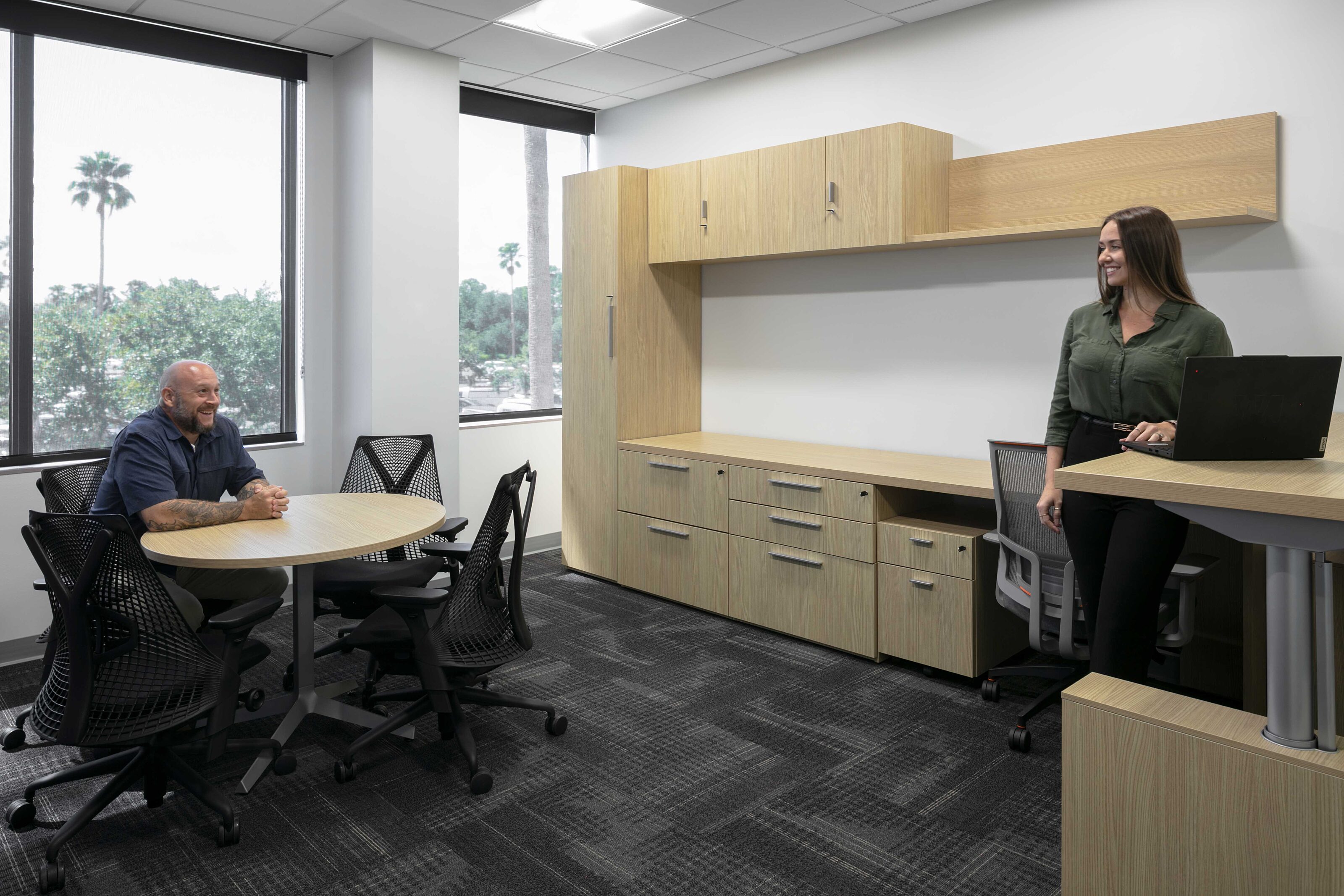 Two employees collaborate in a private office with light wood furniture and meeting seating.