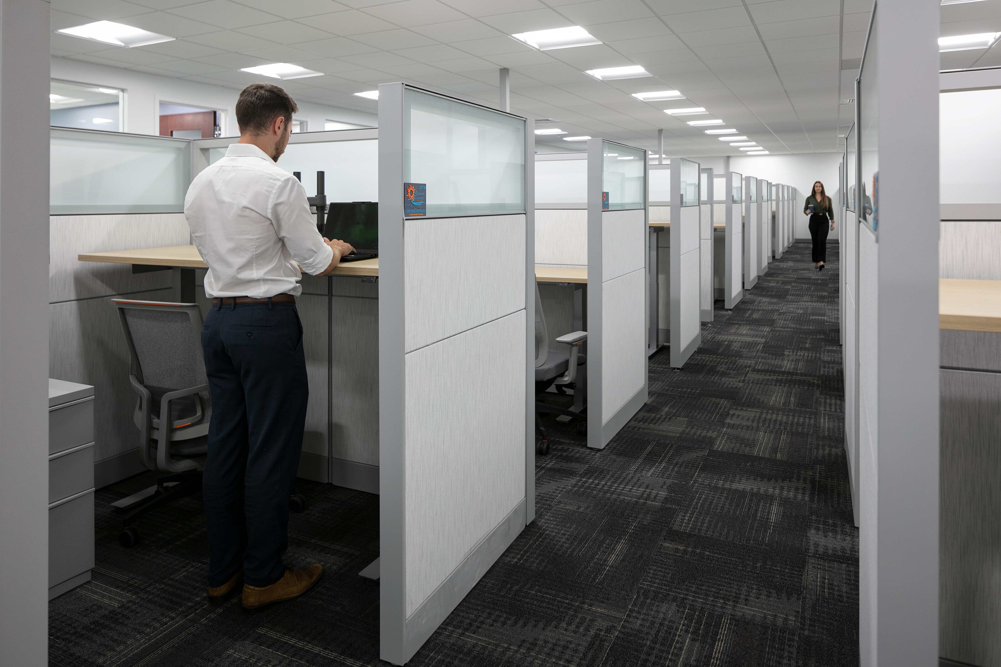 Employee working at a workstation in a modern open office with rows of glass-panel cubicles.