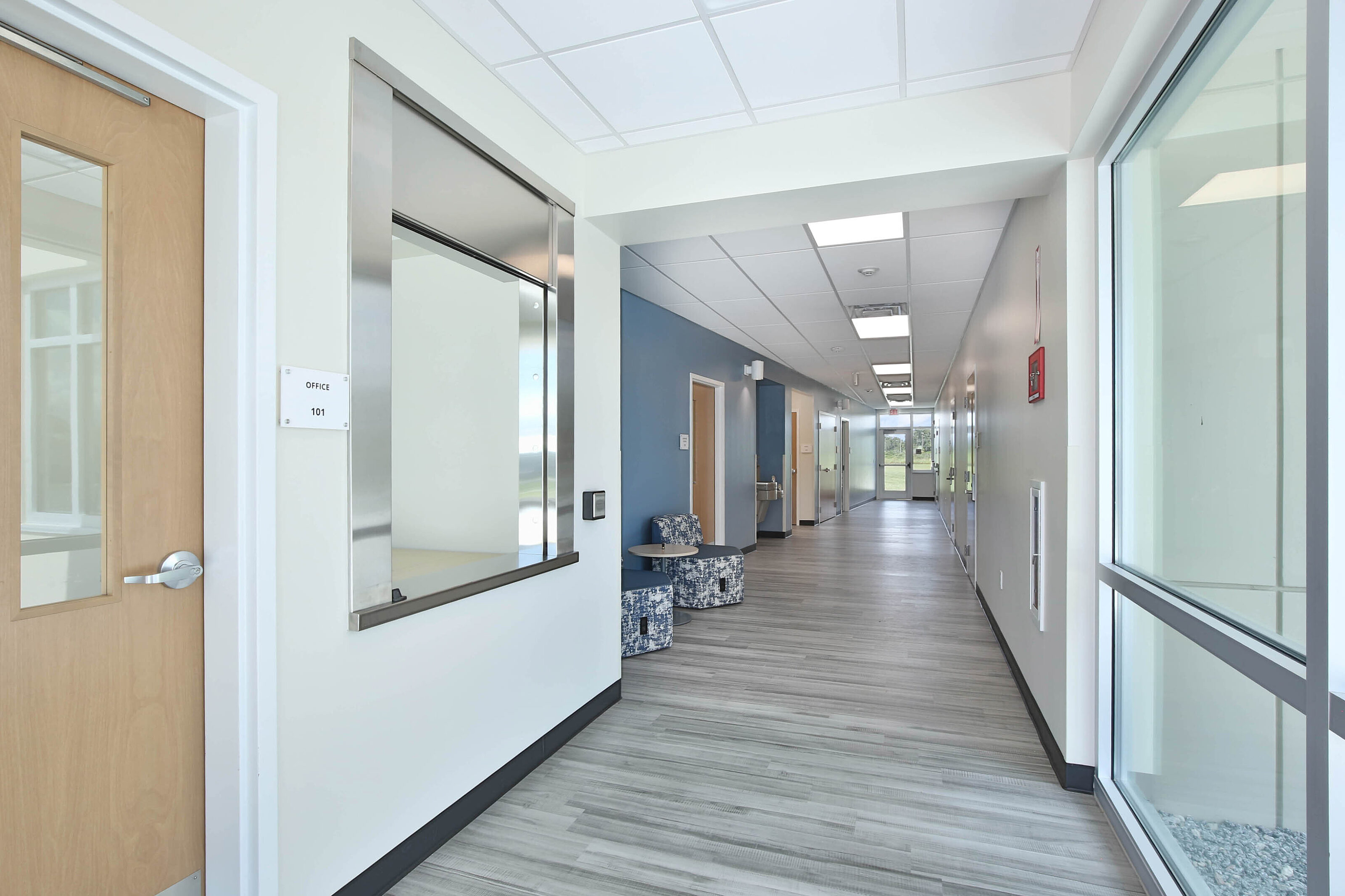 Bright modern hallway with glass windows, wood doors, gray flooring and blue patterned lounge seating.