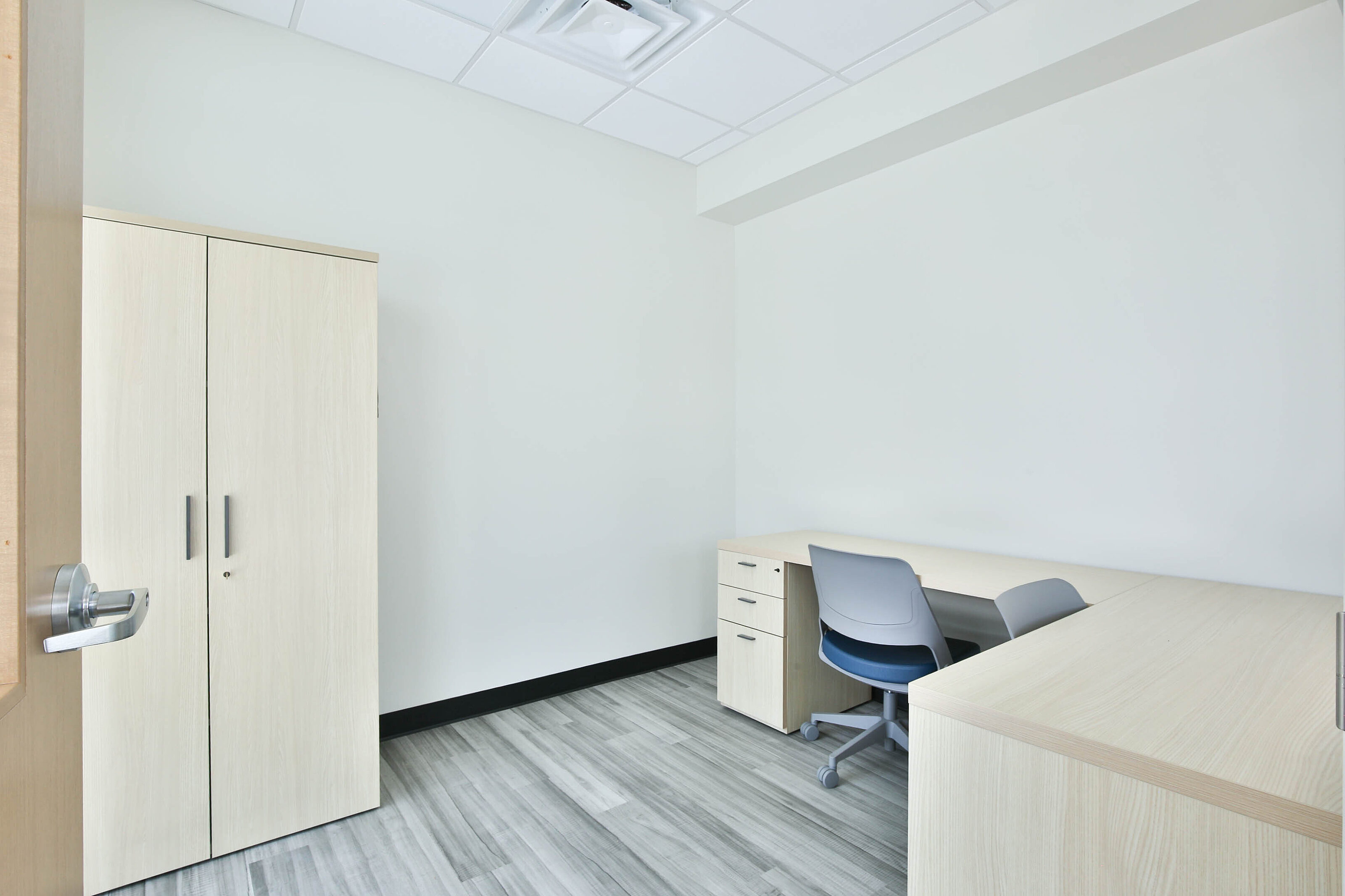 Private office with light wood desks and storage cabinet, gray task chairs, and gray wood-look flooring.