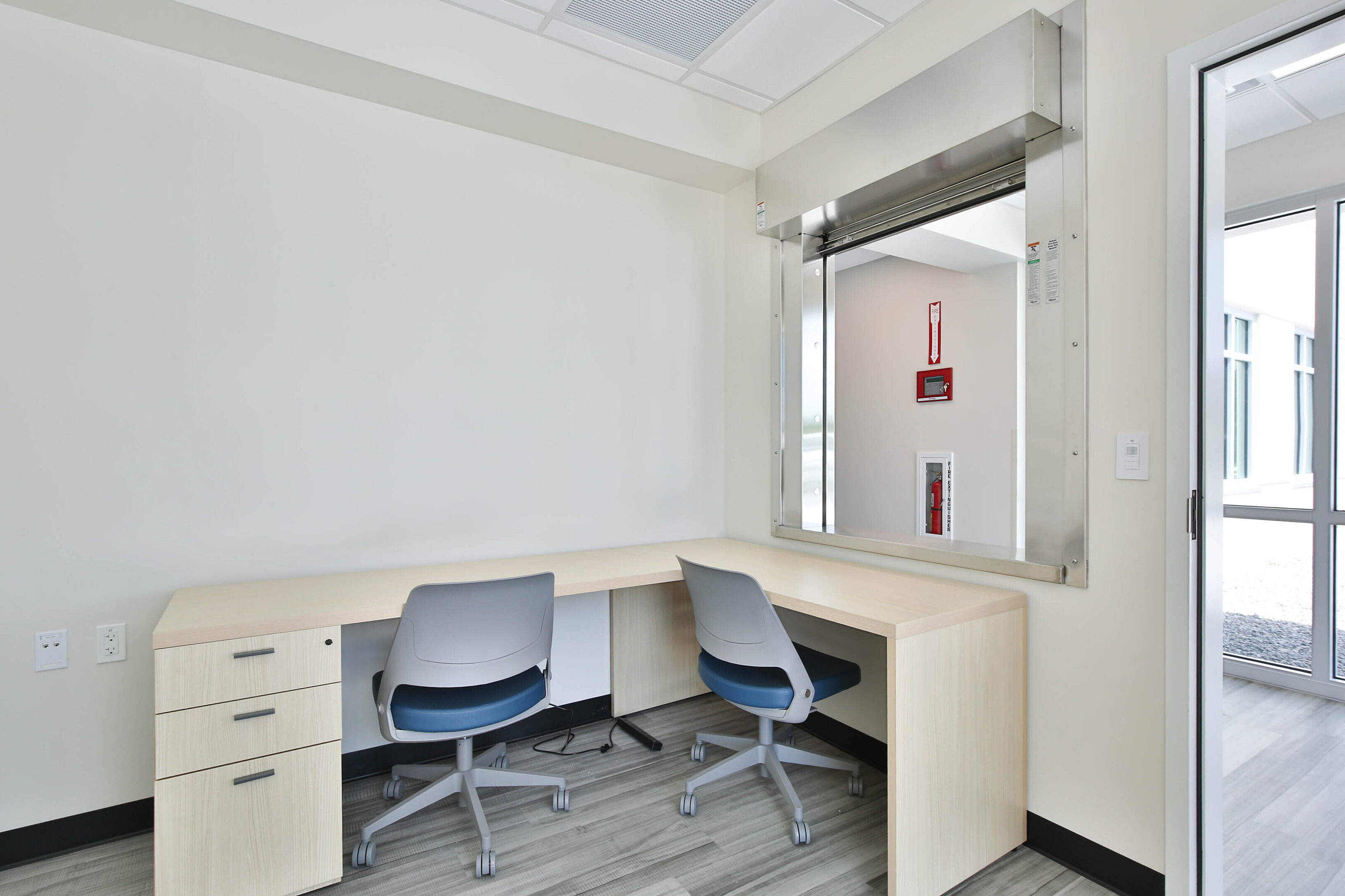 Office workspace with two light wood desks, gray task chairs, a service window, and gray wood-look flooring.