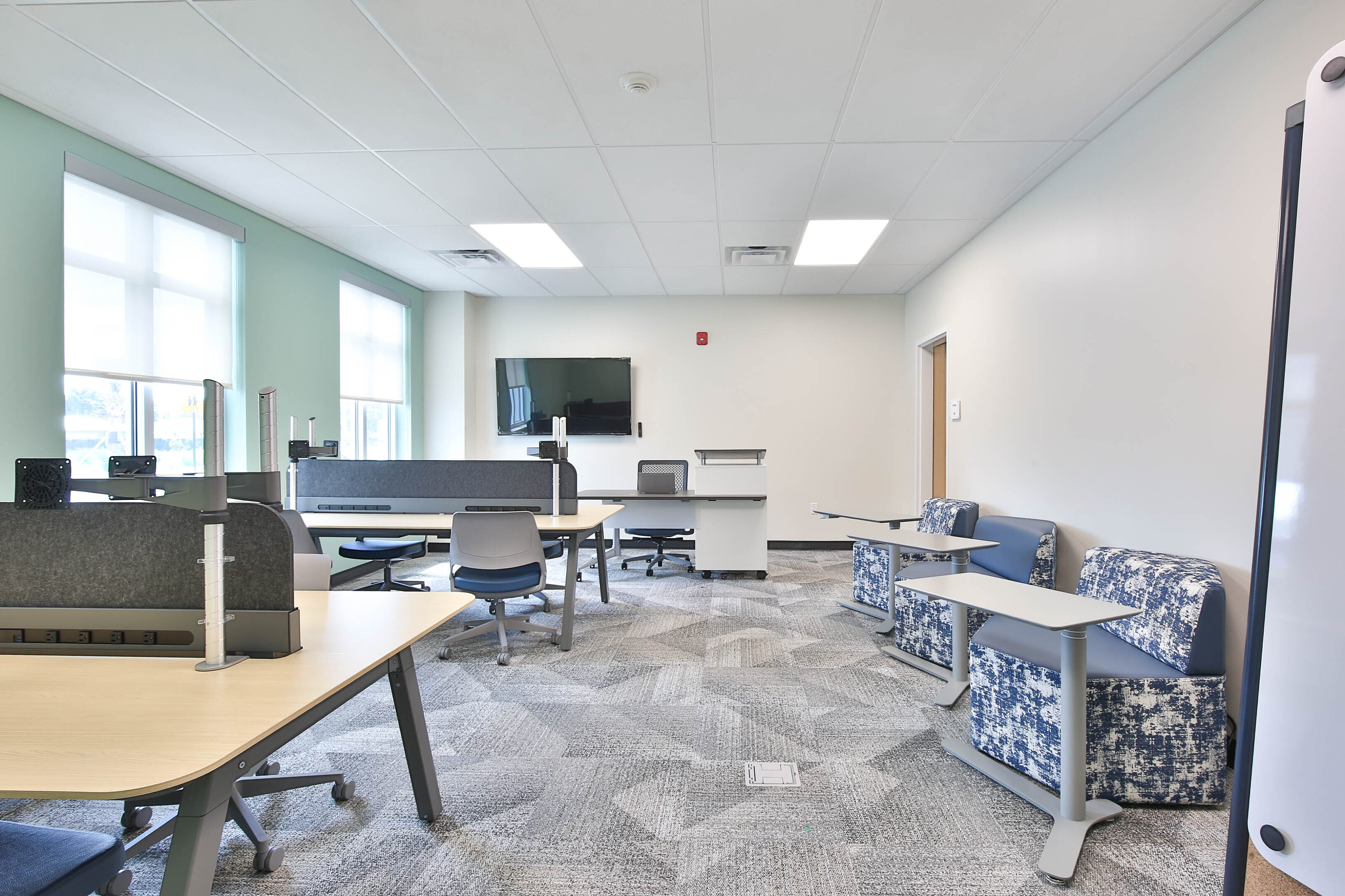 Modern collaborative workspace with shared desks, gray task chairs, blue lounge seating, and patterned carpet.