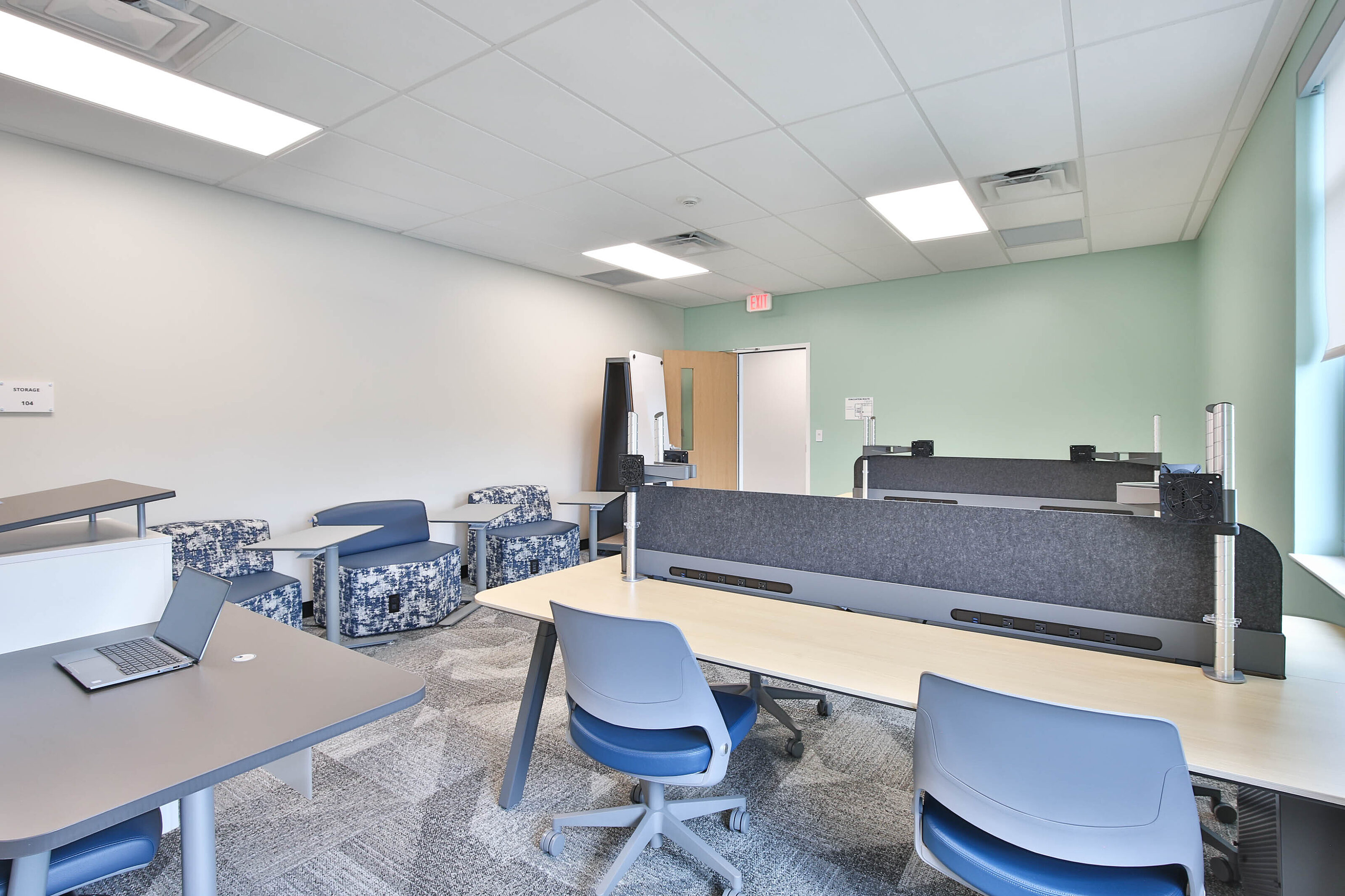 Modern collaborative workspace with shared desks, blue task chairs, patterned lounge seating, and gray carpet.